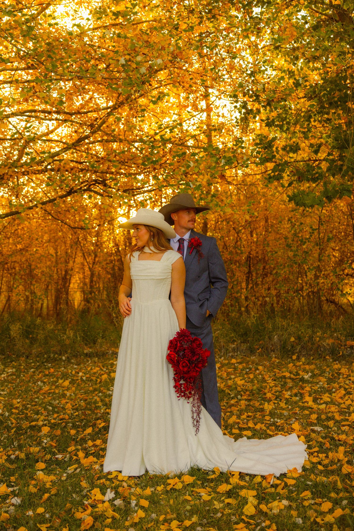 Bride and groom in cowboy hats pose among golden autumn trees, she holds red bouquet, wearing white gown.