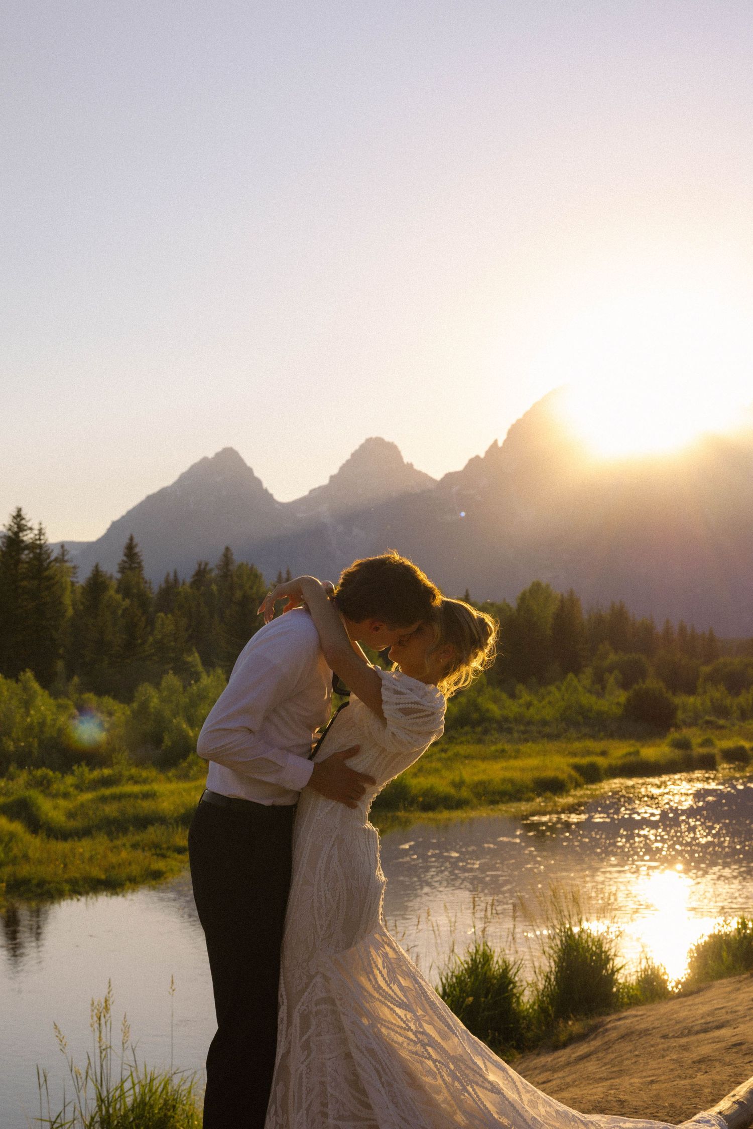Bride and groom embrace at sunset with Grand Teton mountains and reflective lake in background during outdoor wedding.