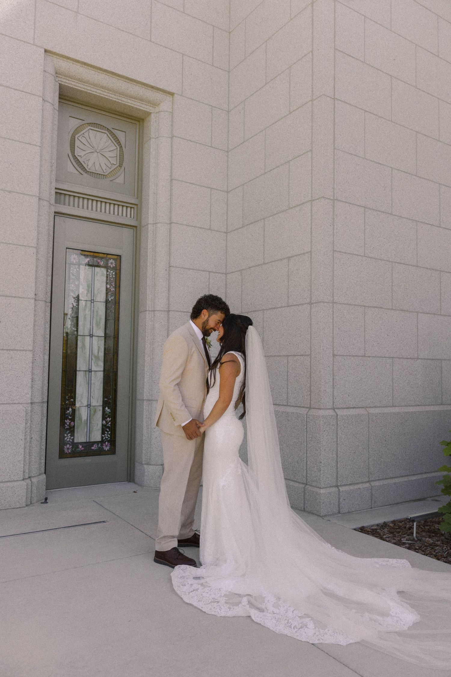 Bride and groom share a romantic moment outside a white stone temple, her cathedral veil trailing elegantly.