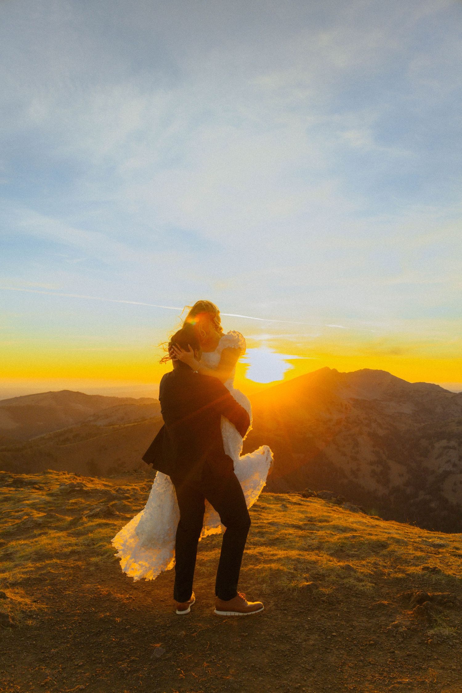 Couple embracing at golden sunset on mountain peak, groom lifting bride in wedding attire with vibrant orange sky backdrop.