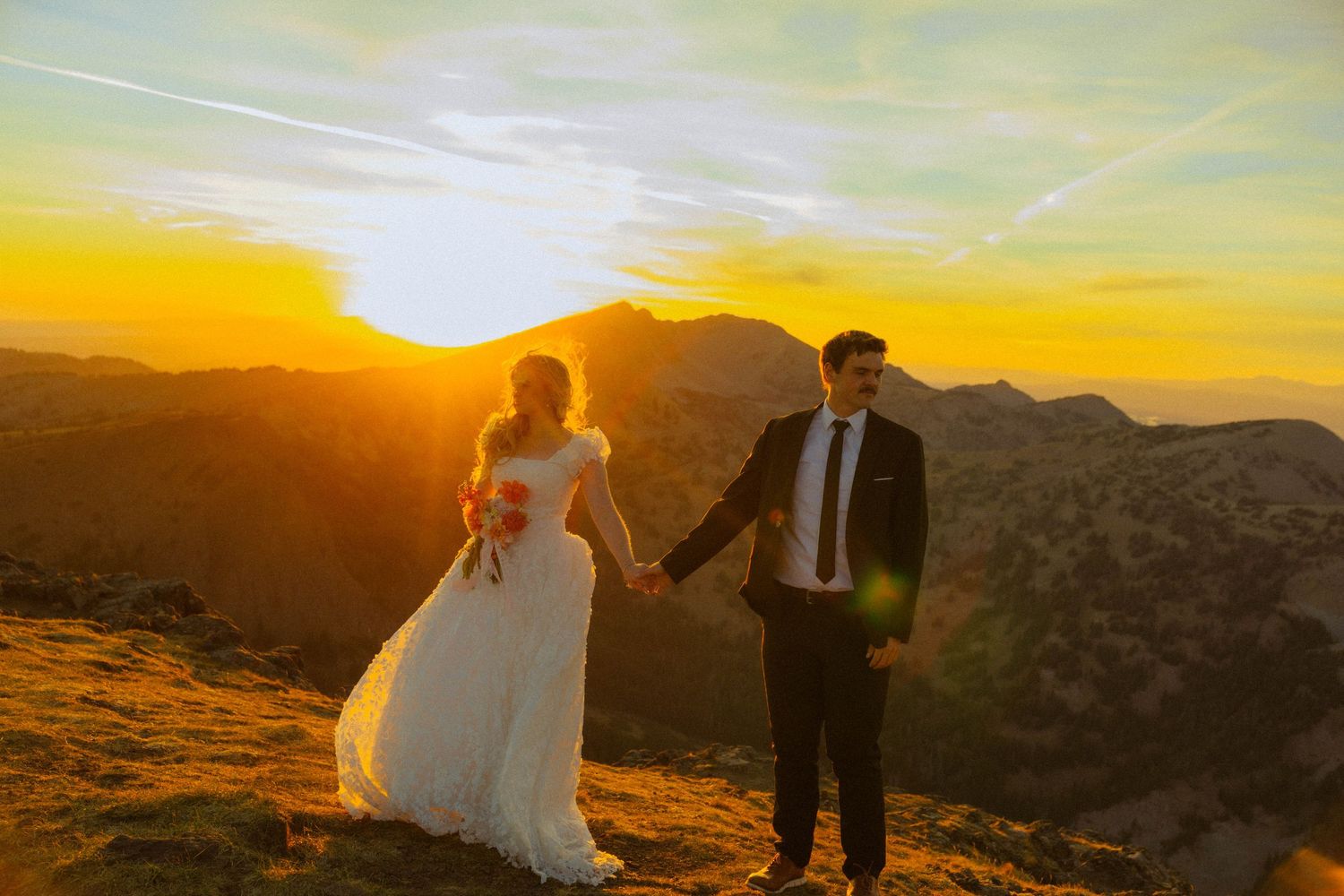 Bride and groom holding hands at golden sunset on mountain peak in wedding attire.