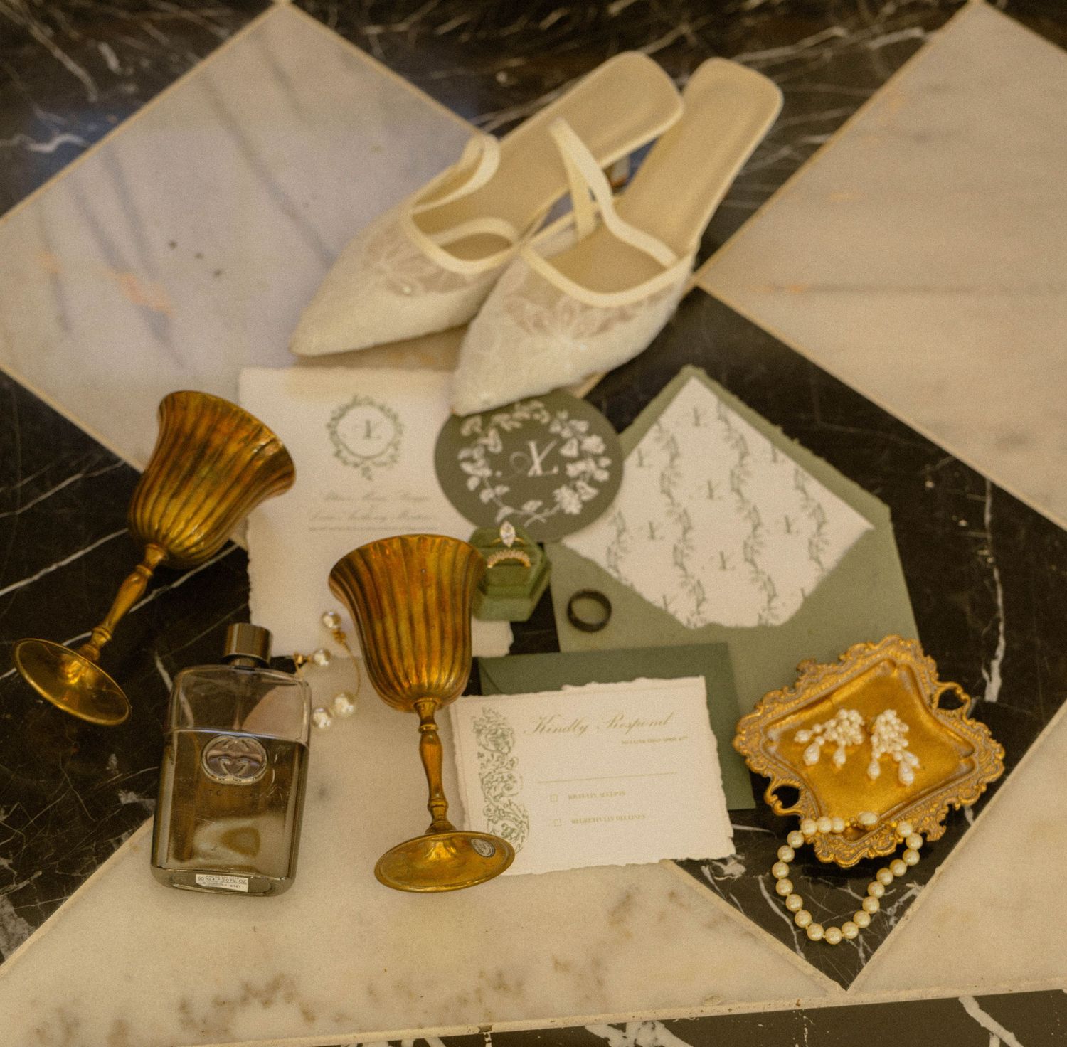 Elegant wedding flat lay with gold goblets, white heels, pearl jewelry, perfume, and lace invitation on marble.