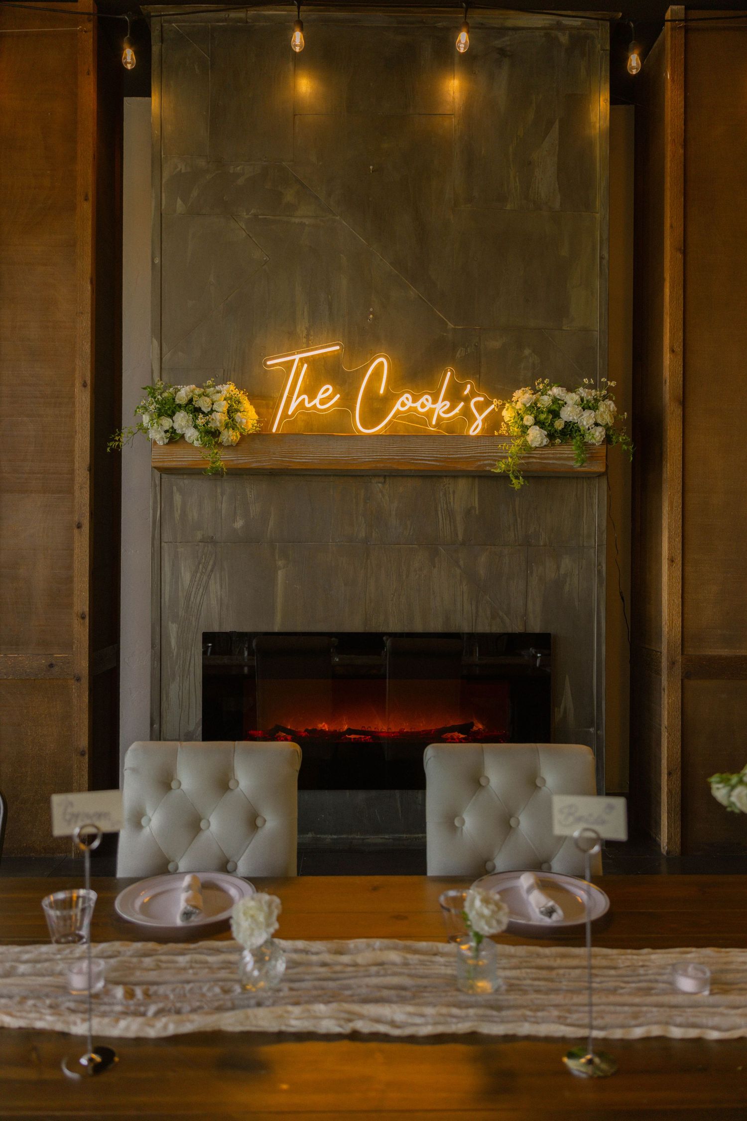 Elegant wedding sweetheart table with tufted chairs, glowing neon sign reading The Cooks above a fireplace with floral arrangements.