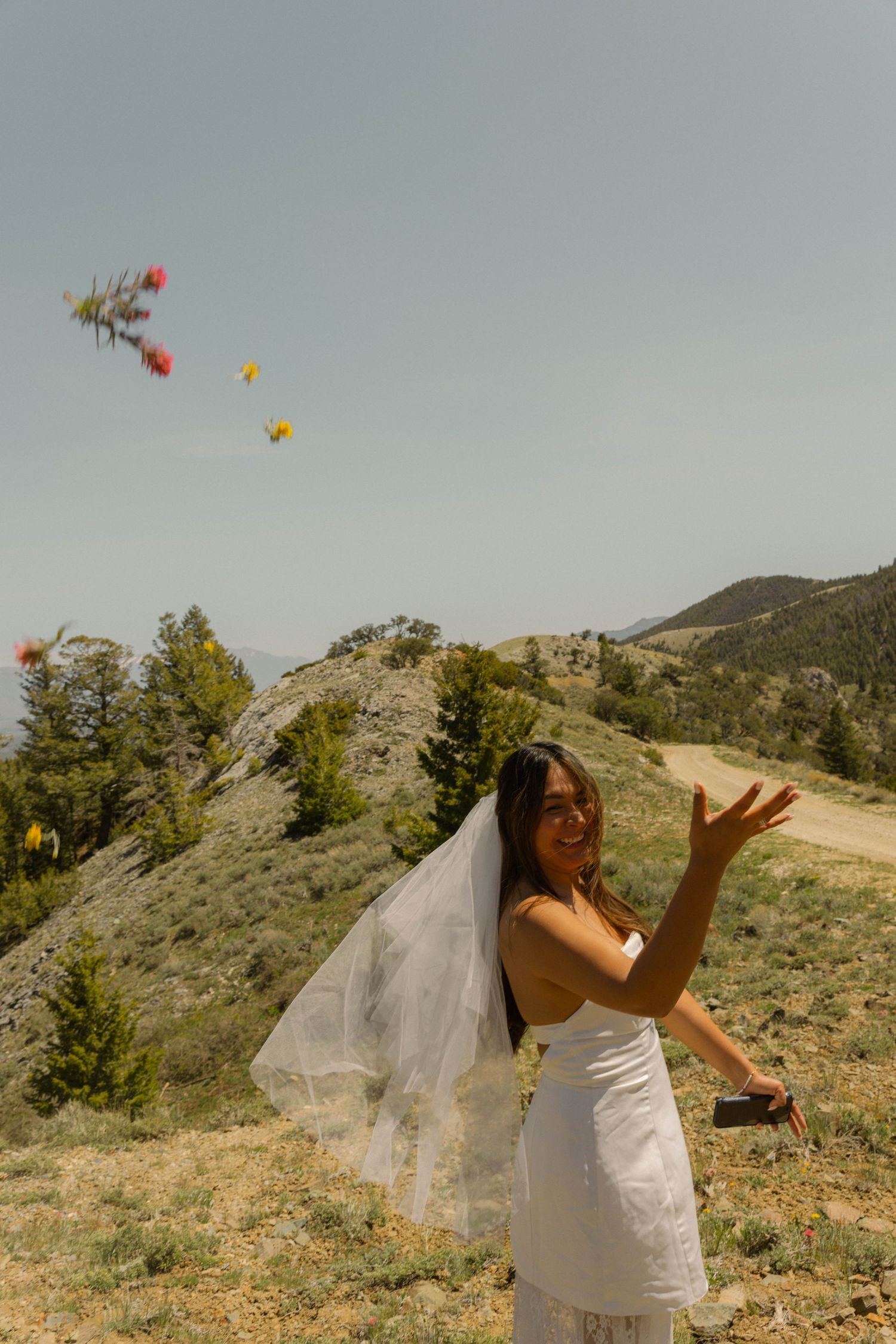 Bride in white dress and veil tossing bouquet outdoors with mountain landscape backdrop on sunny day.