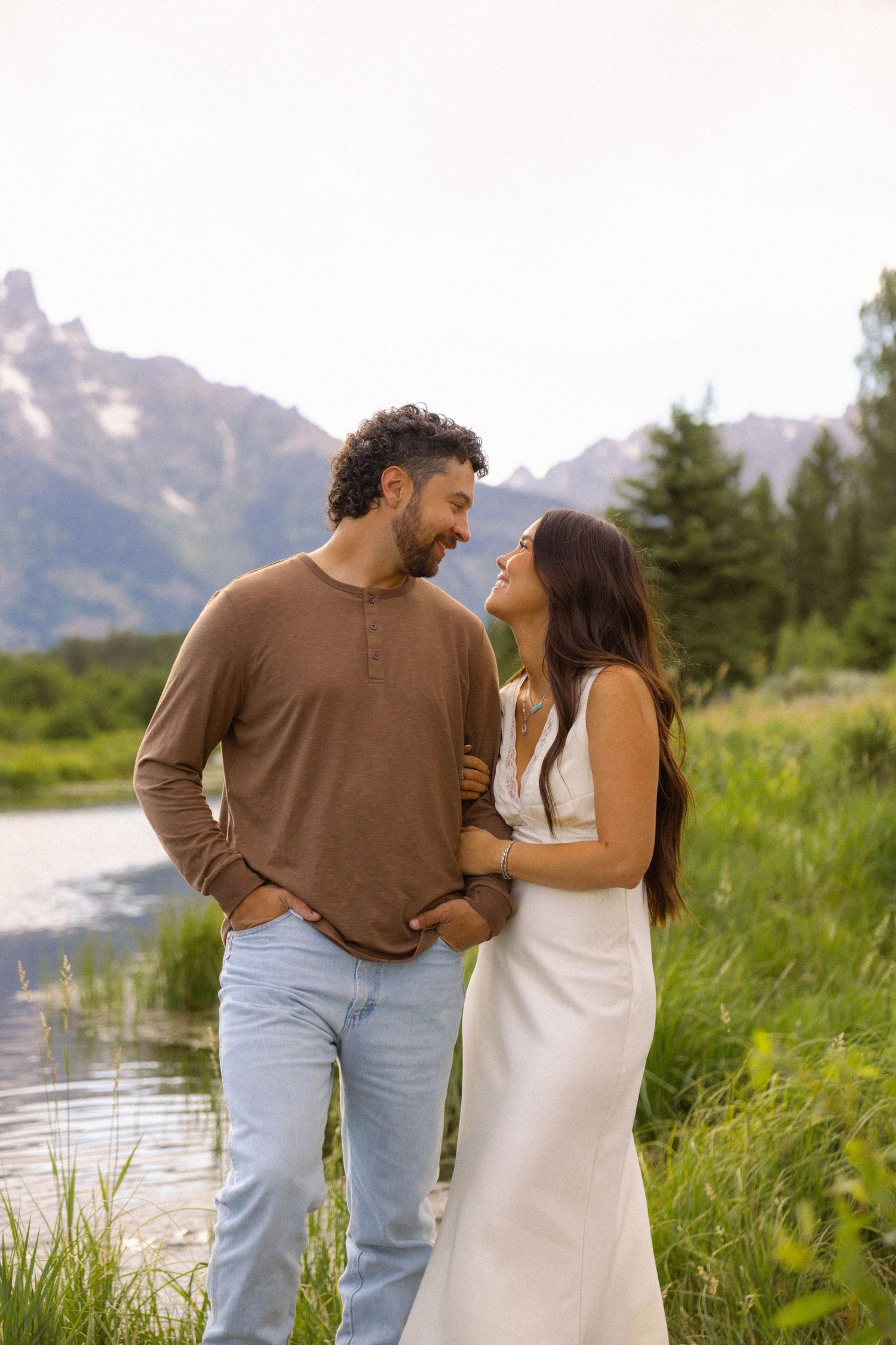 Couple smiling at each other in white dress and brown shirt by mountain lake in Grand Teton National Park.
