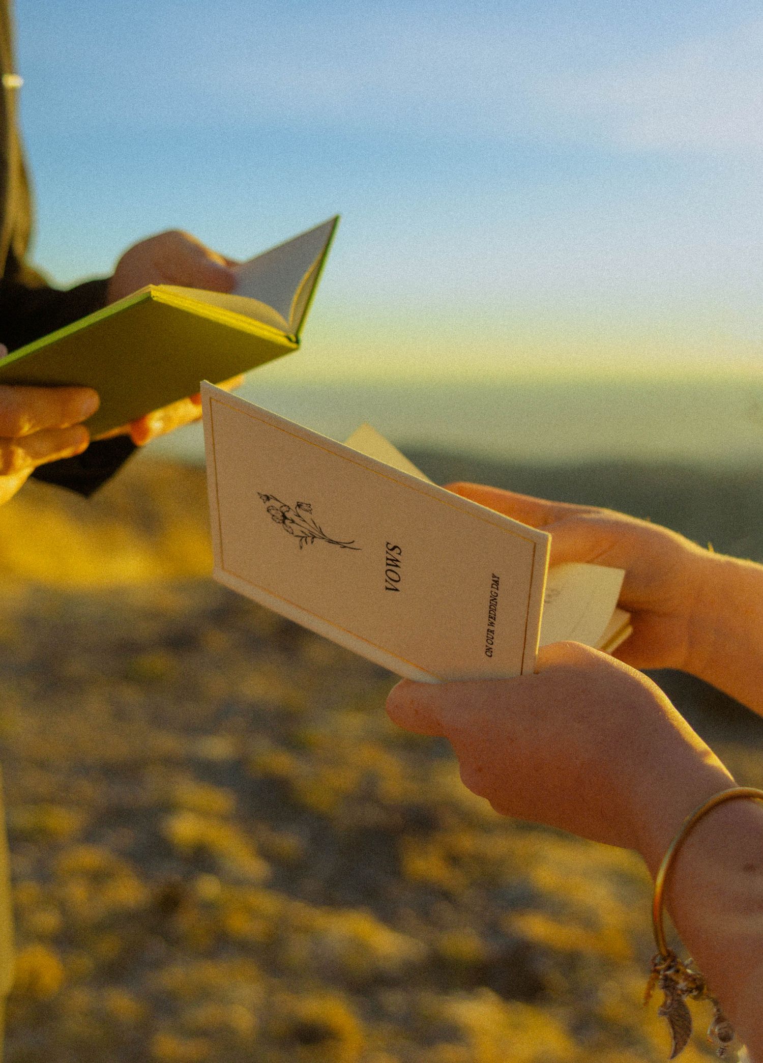Two people exchange wedding vows booklets outdoors at golden hour with scenic landscape in background.