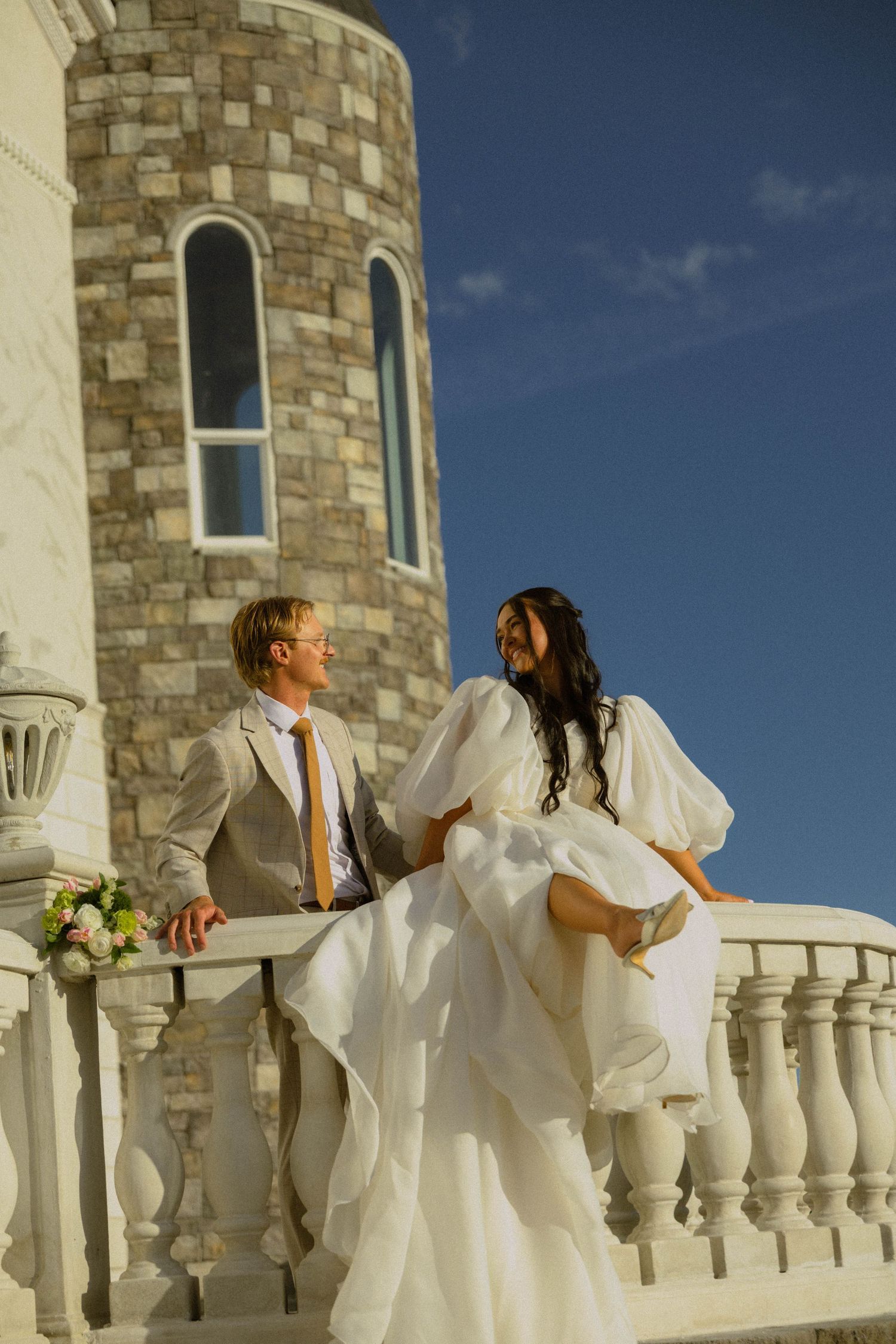 Bride in white gown and groom in tan suit pose on a sunlit balcony beside a stone tower on their wedding day.