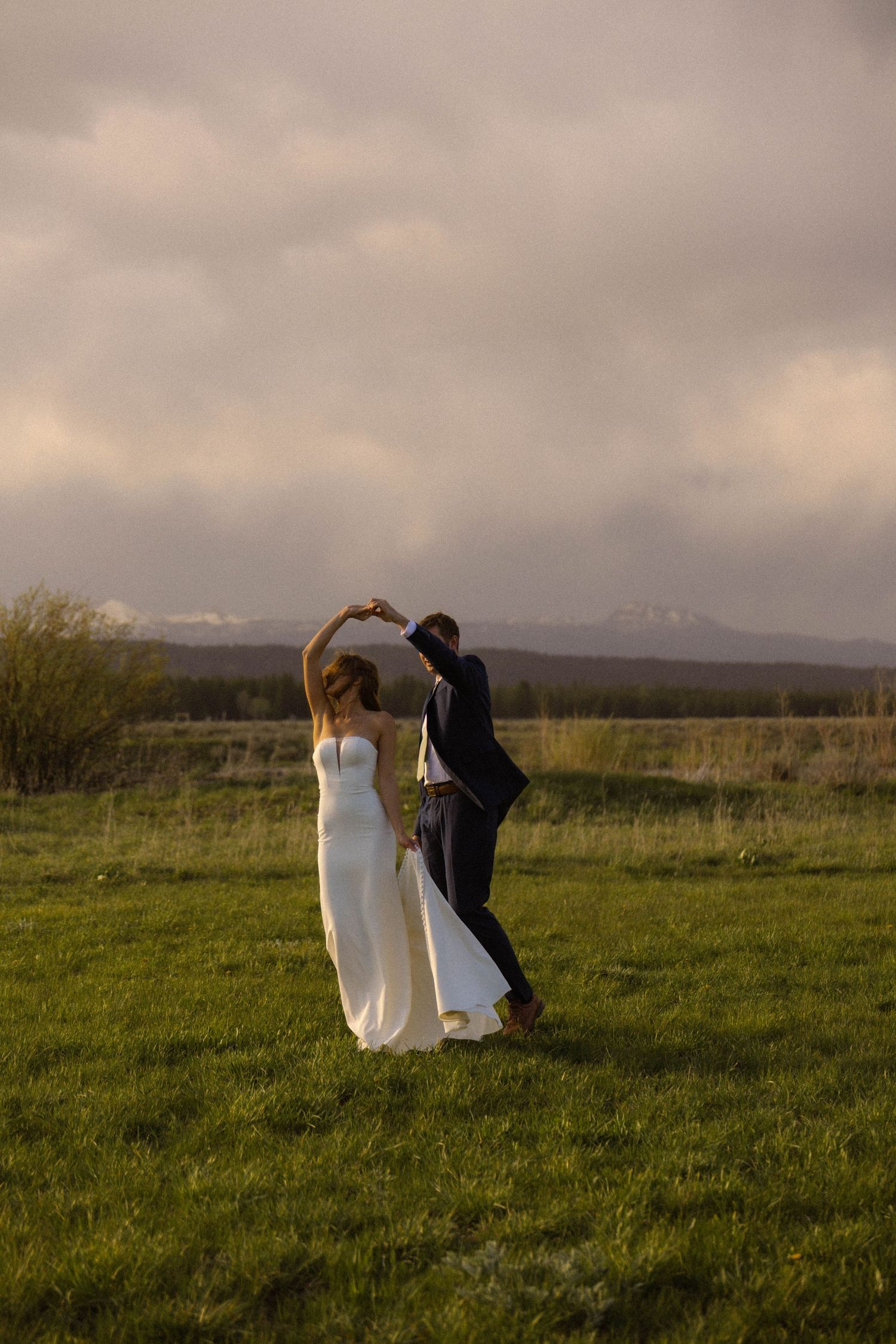 Bride and groom dancing in a green field with mountain backdrop during golden hour at outdoor wedding.