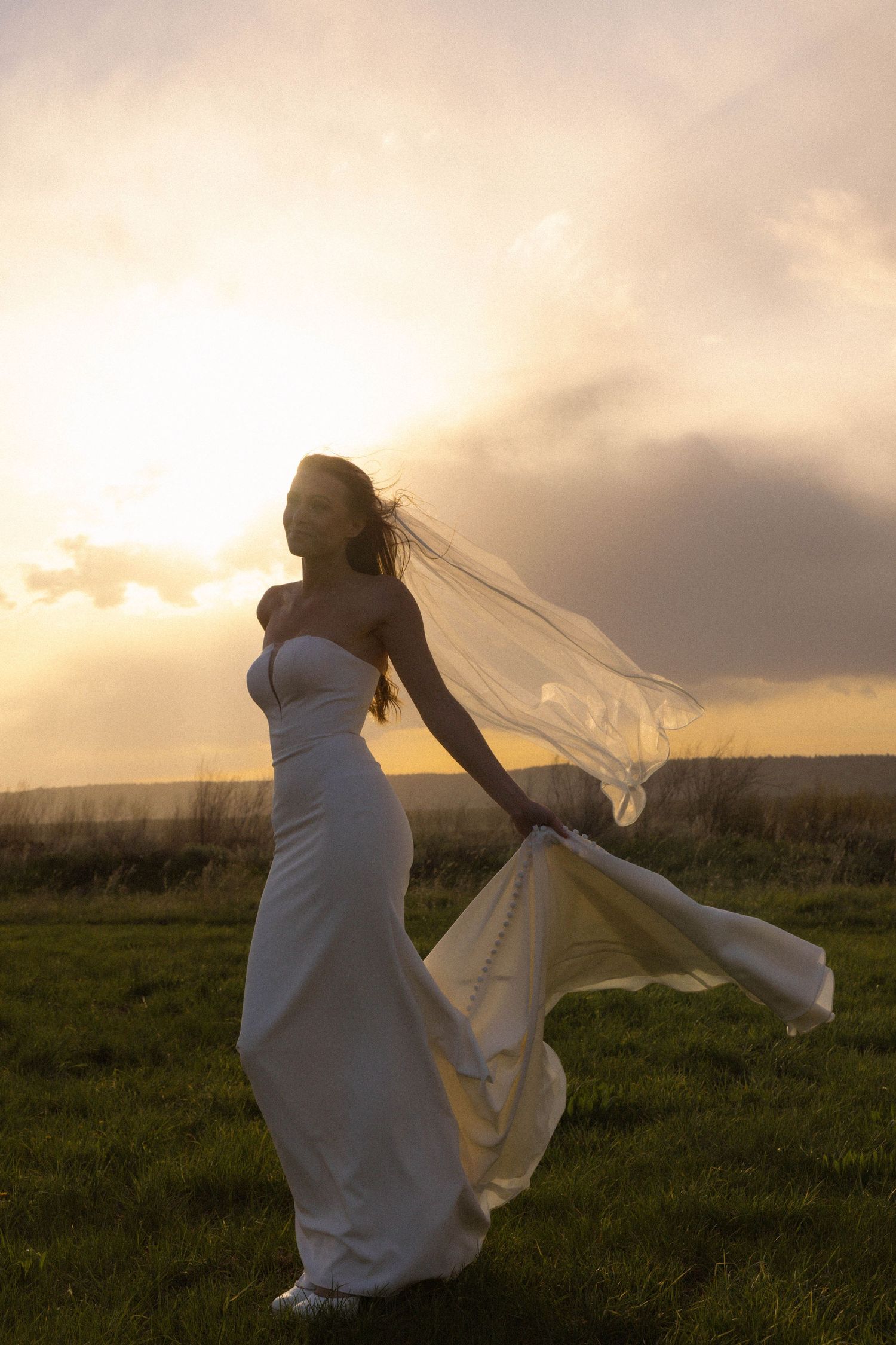 Bride in white gown with flowing veil standing in green field at golden sunset, hair and dress blowing in wind.