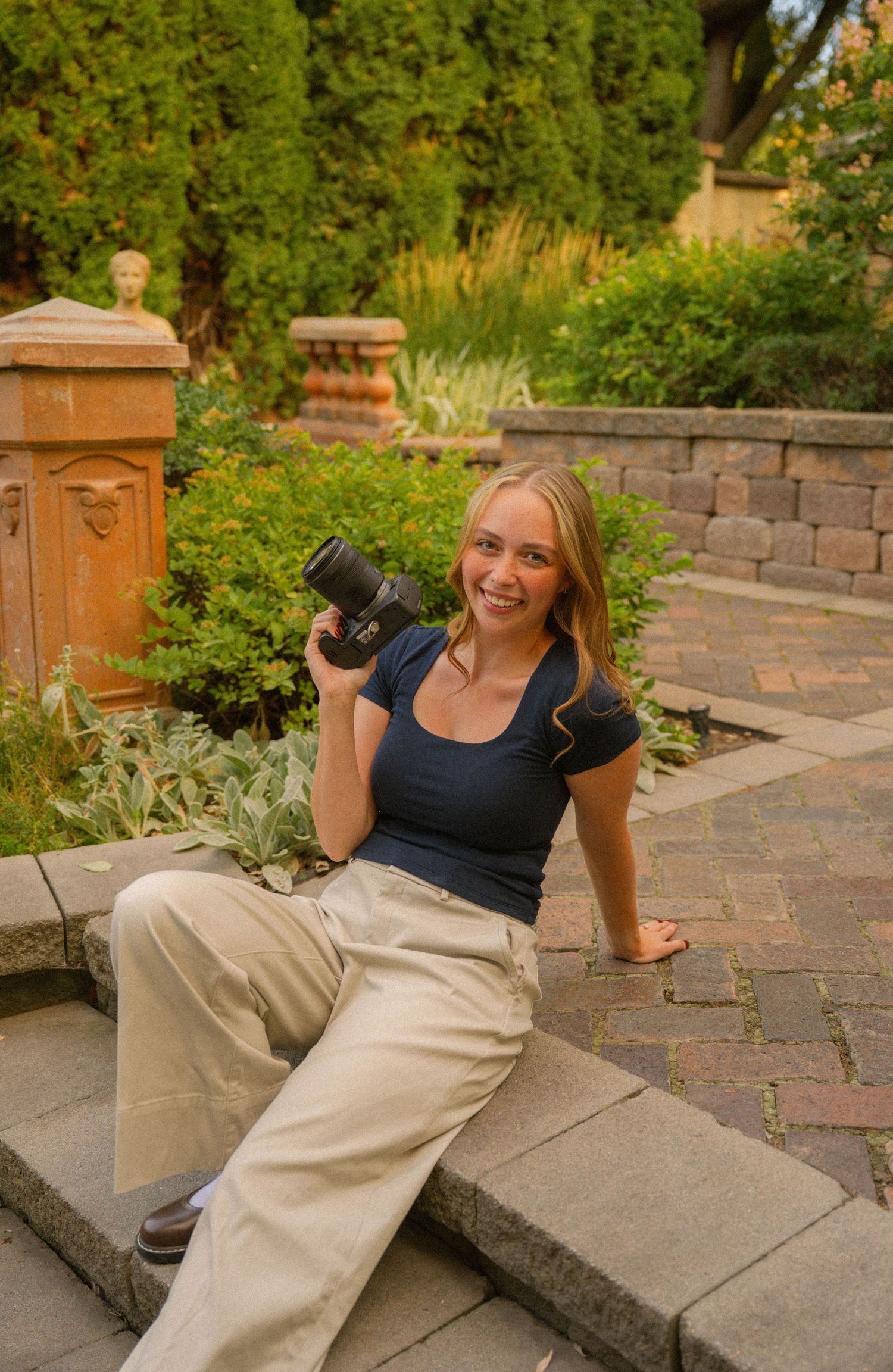 Young blonde woman smiling outdoors on stone steps, holding a camera, wearing navy top and khaki pants, surrounded by lush greenery.