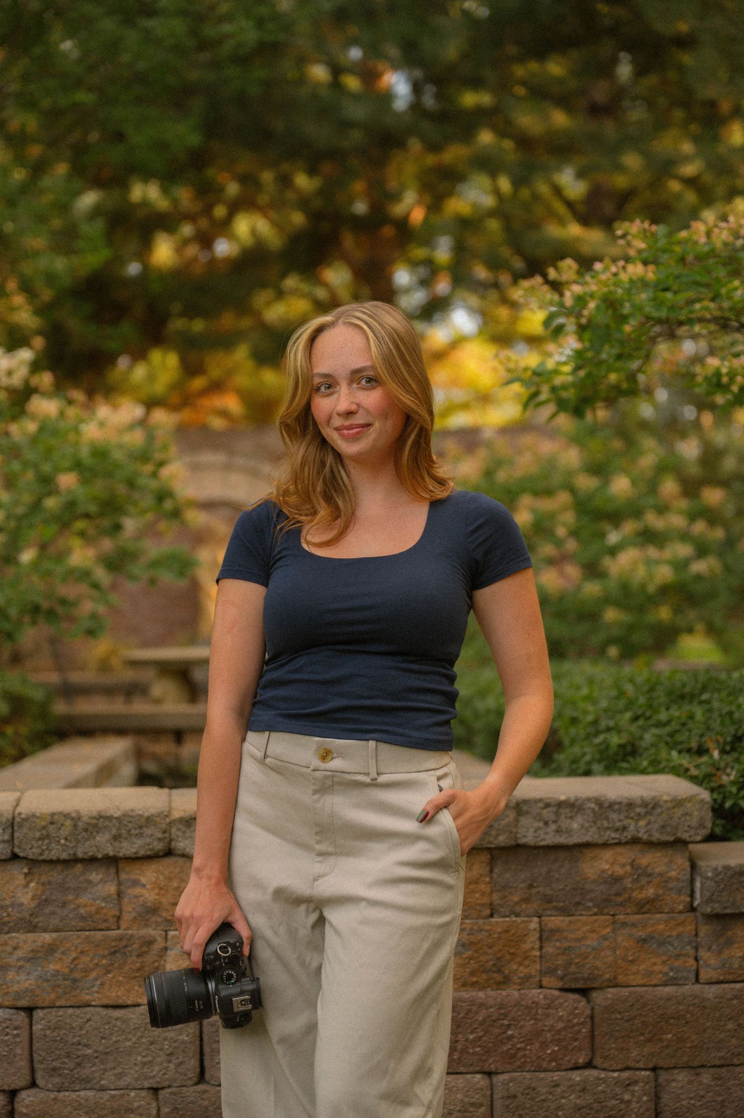 Young blonde woman holding a DSLR camera, wearing navy top and khaki pants, standing outdoors near a stone wall.