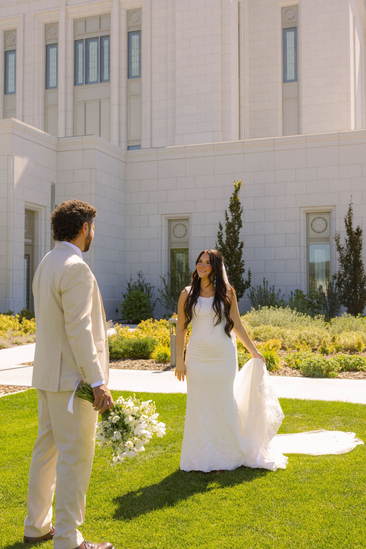 Bride in fitted white gown smiles at groom holding bouquet on lush green lawn outside white temple.