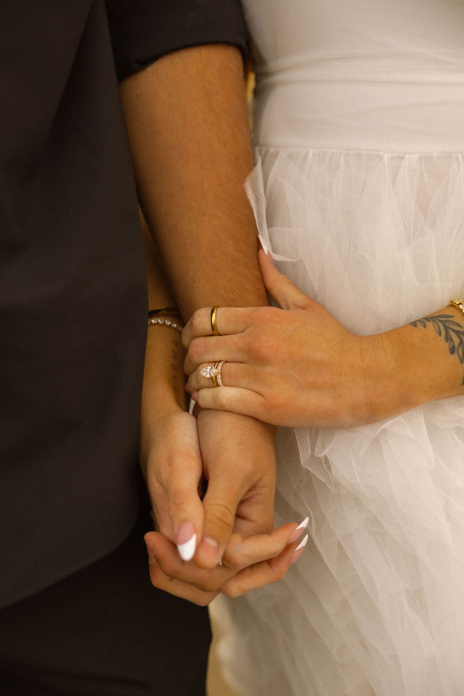 Bride and groom holding hands, showcasing wedding rings, diamond engagement ring, and floral wrist tattoo in white tulle gown.