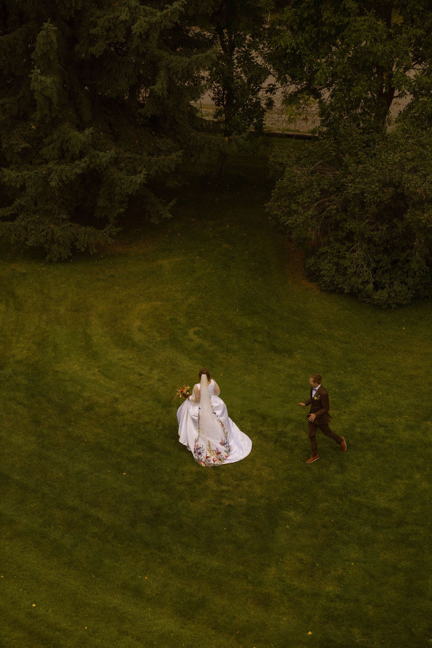 Aerial view of bride in white gown and groom in dark suit walking across lush green lawn surrounded by trees.
