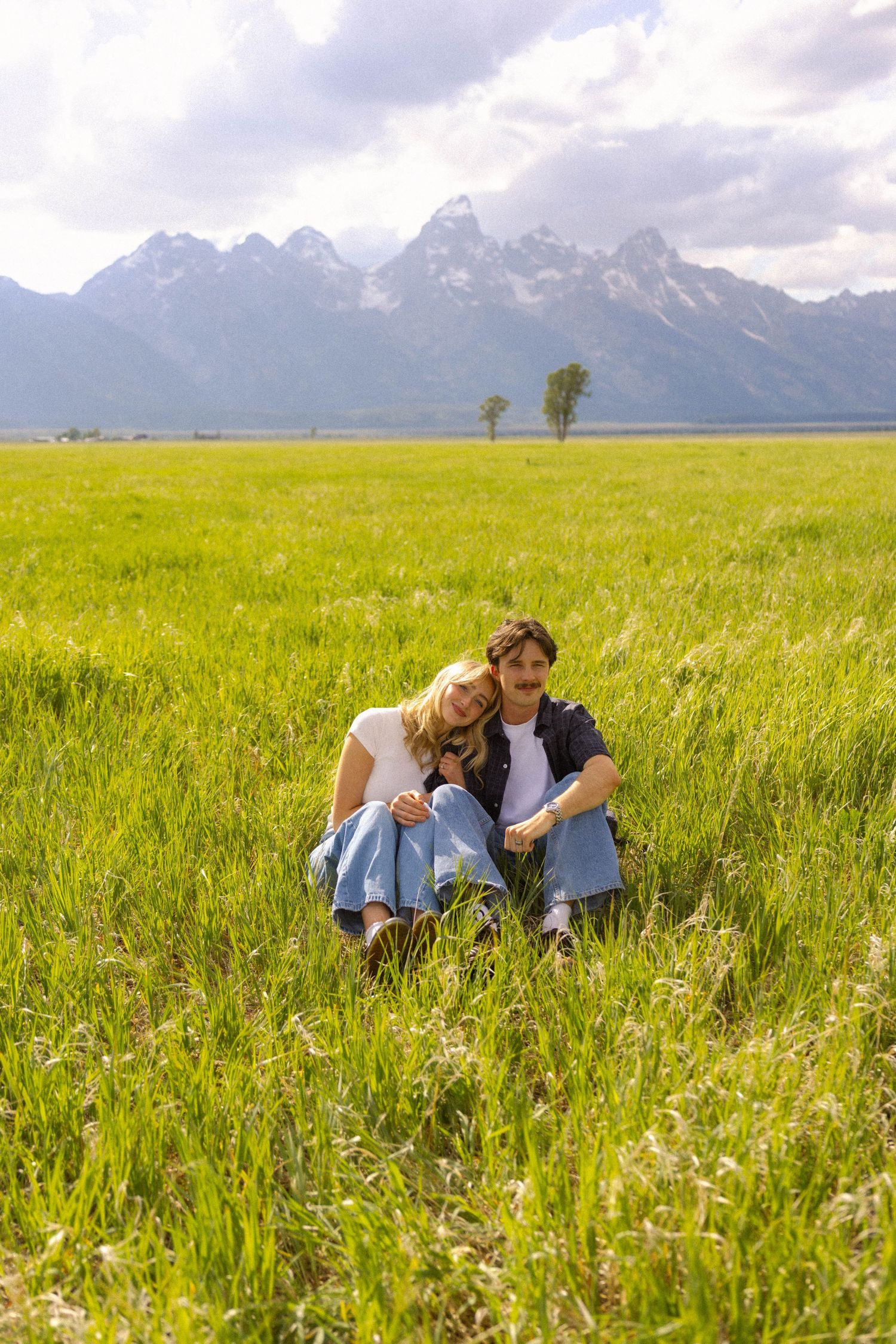 Couple sitting together in a lush green meadow with the Grand Teton mountain range in the background.