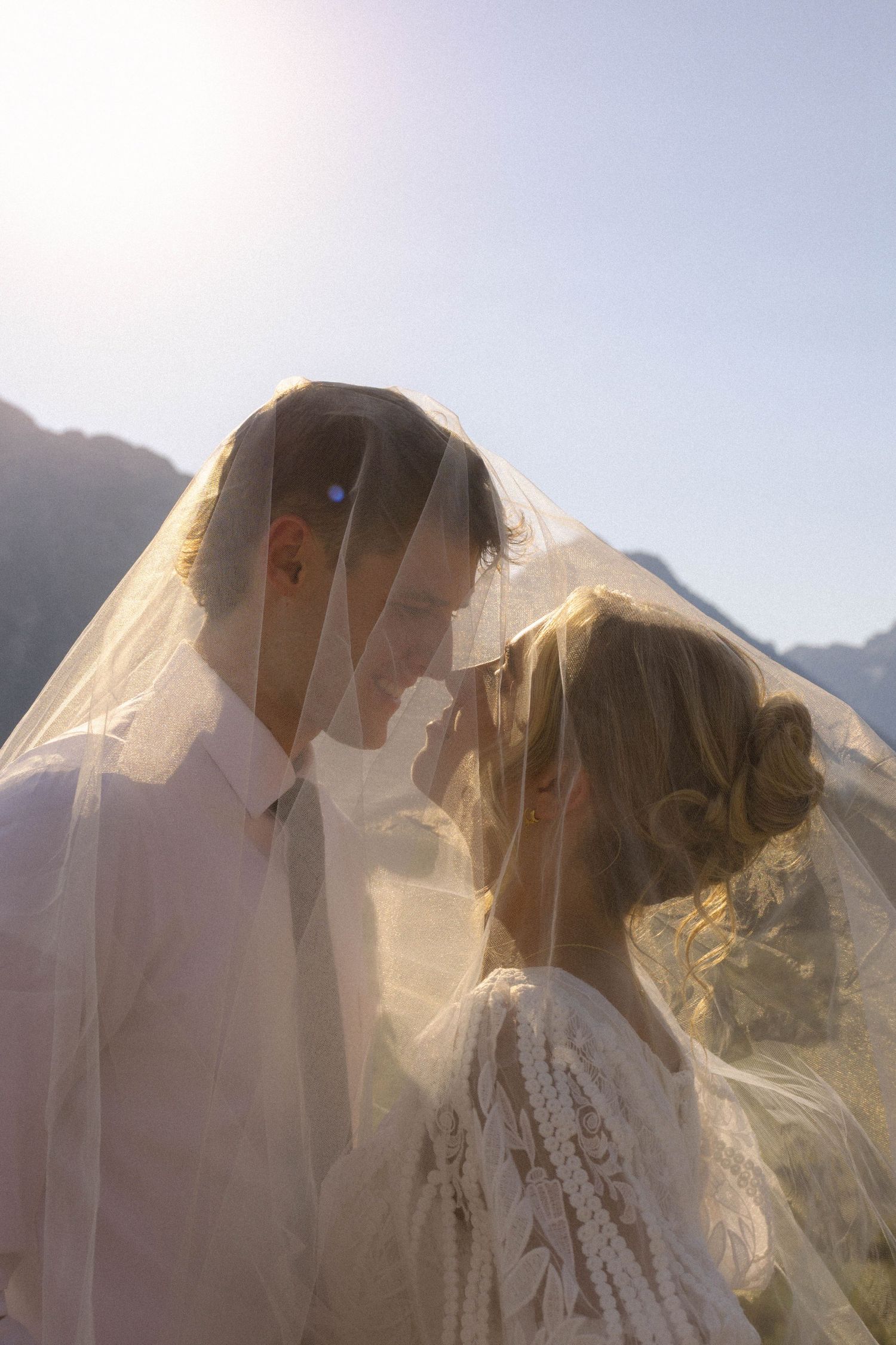 Bride and groom sharing romantic moment under wedding veil with sunlit mountains in background.