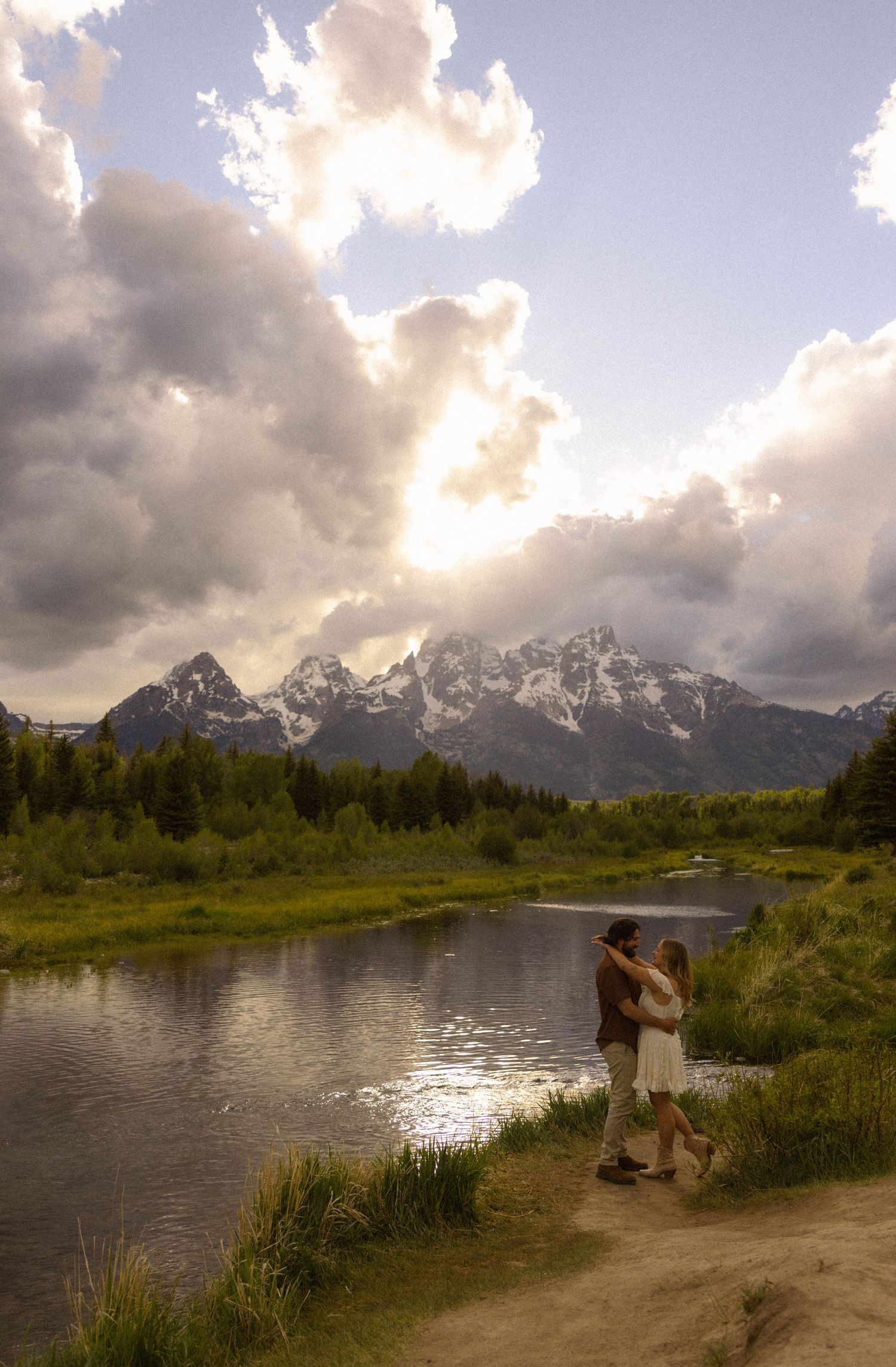 Couple embracing by a river with Grand Teton mountains and dramatic clouds in background at sunset.