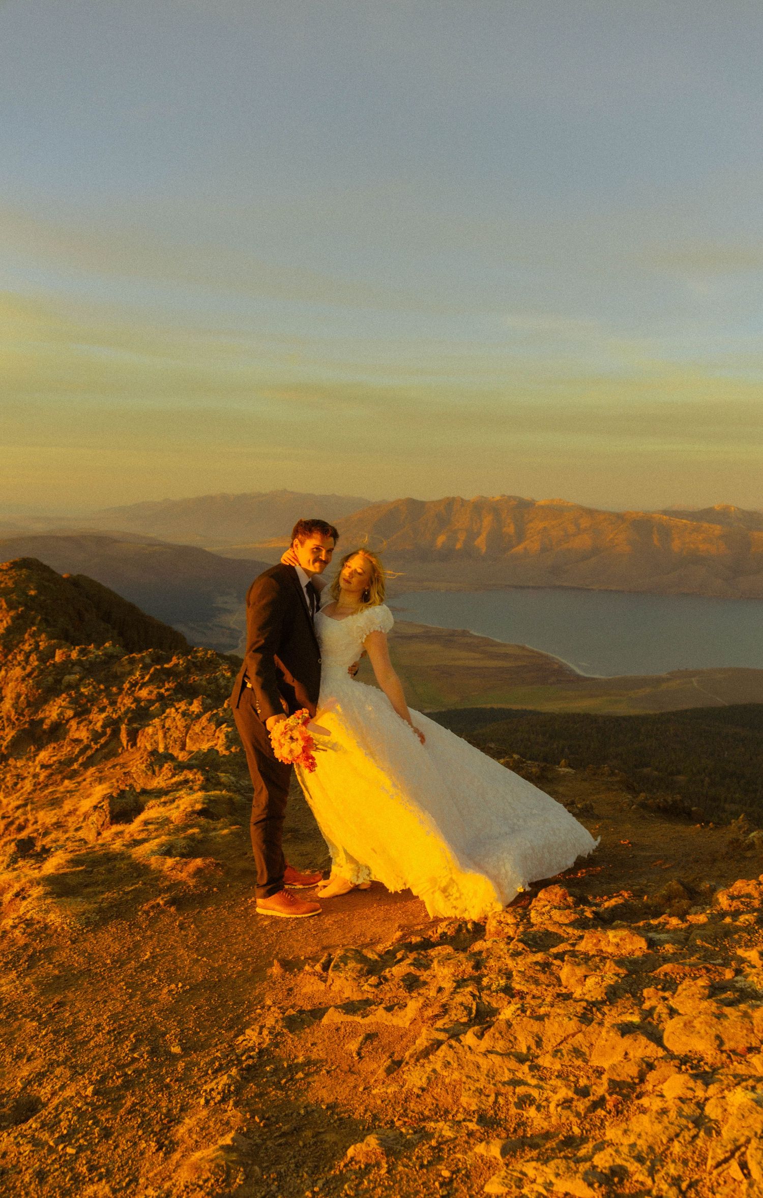 Bride and groom embrace on a mountain summit at golden sunset with scenic lake views.
