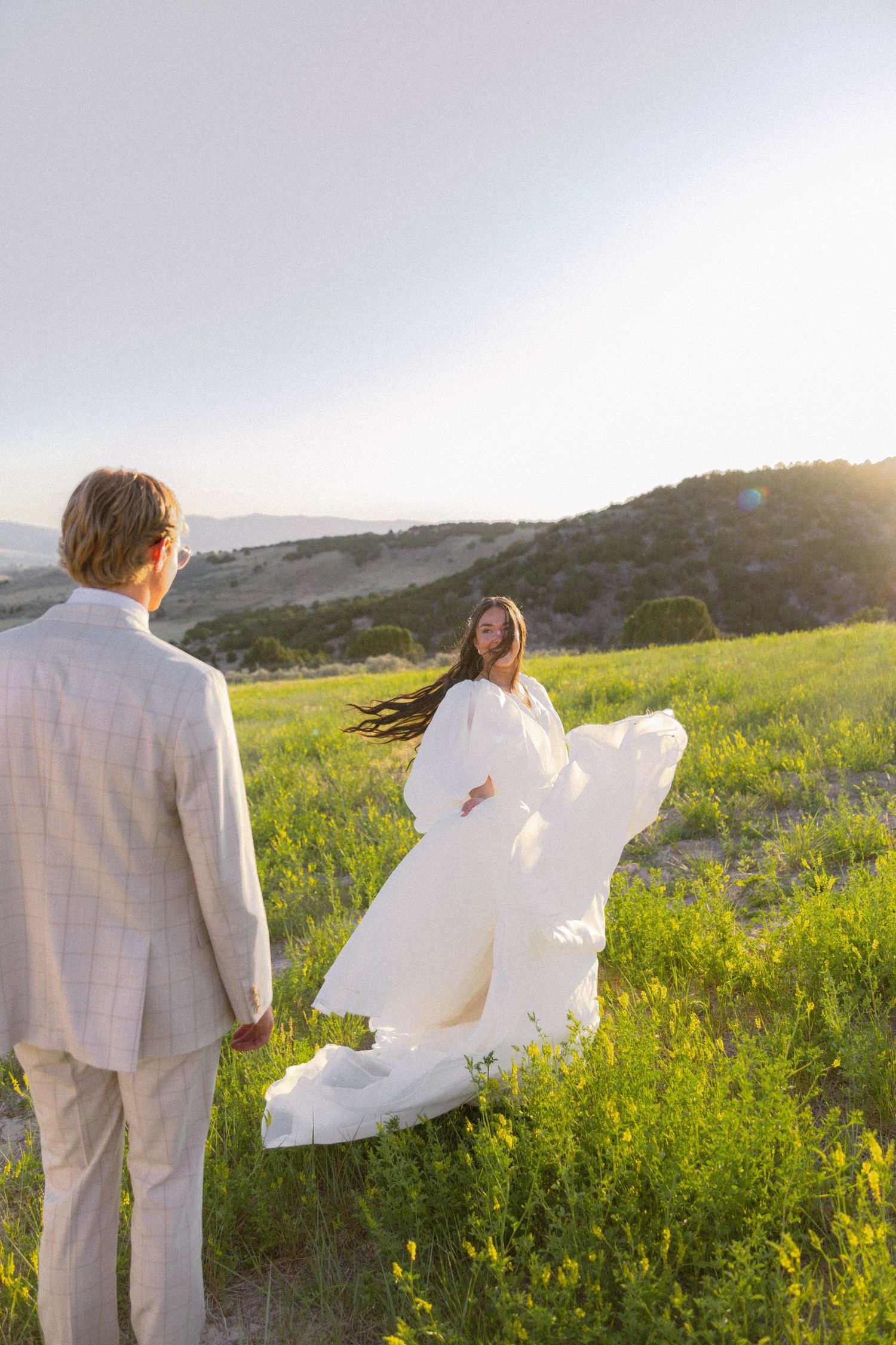 Bride in flowing white gown twirls in green meadow at sunset while groom in tan suit watches.