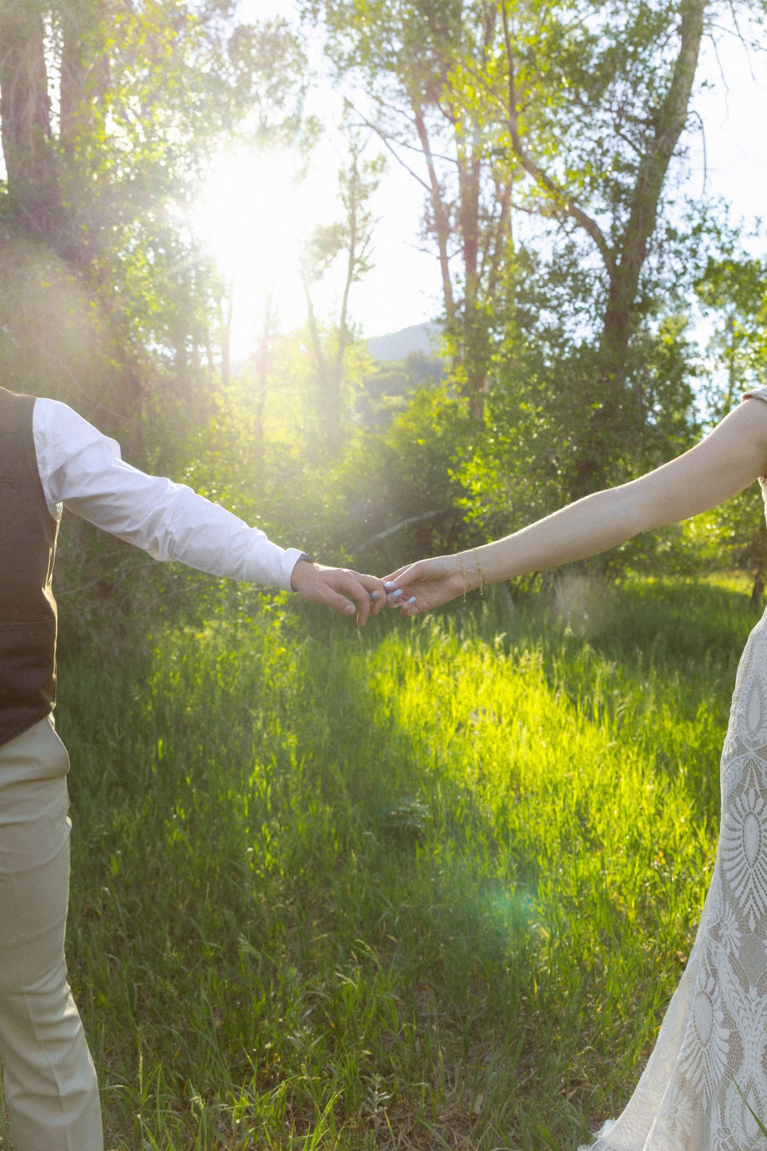 Bride and groom holding hands in a sunlit green meadow surrounded by trees during an outdoor wedding ceremony.
