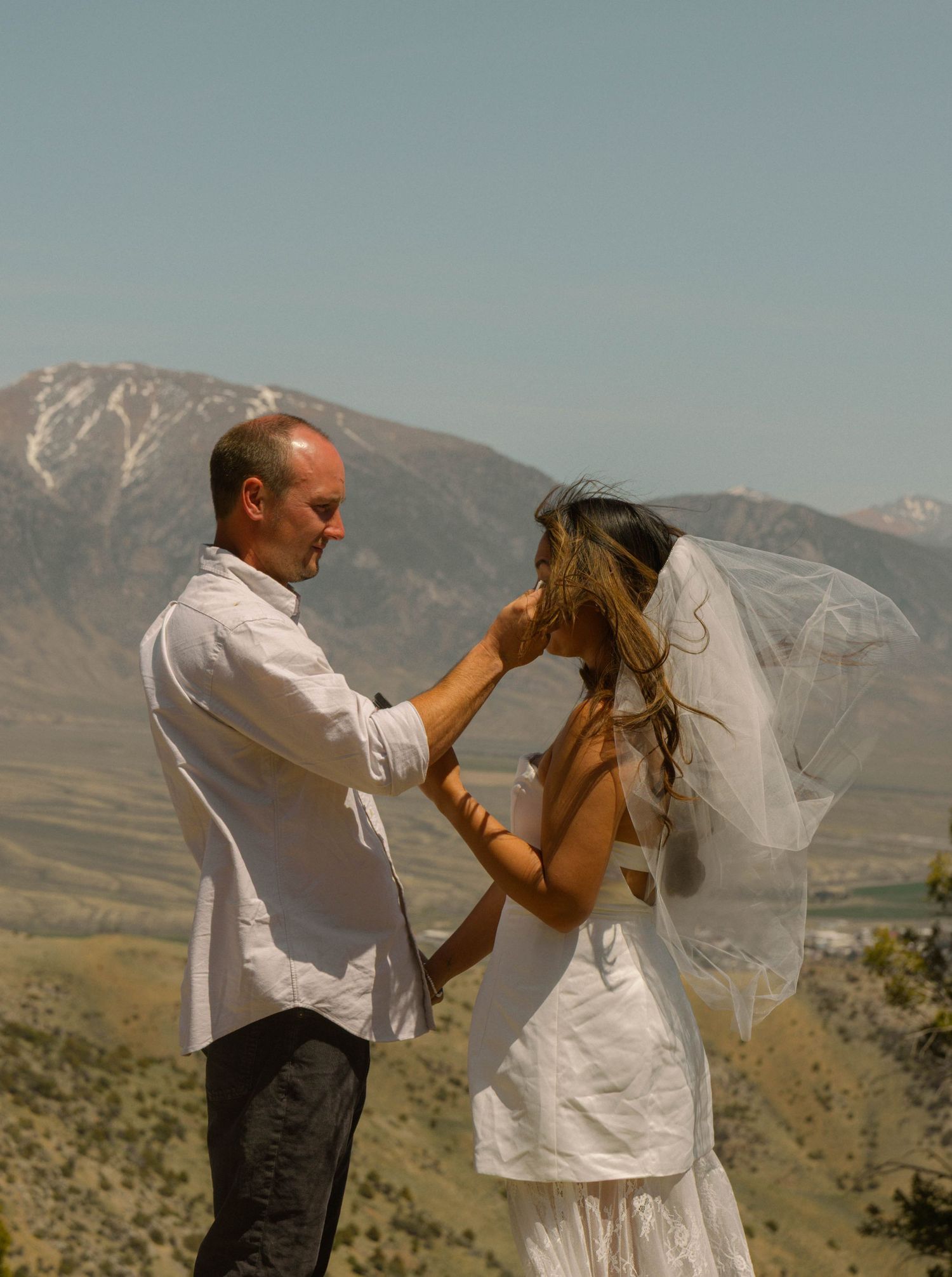 Groom adjusting bride's veil outdoors with scenic mountain landscape backdrop on their wedding day.