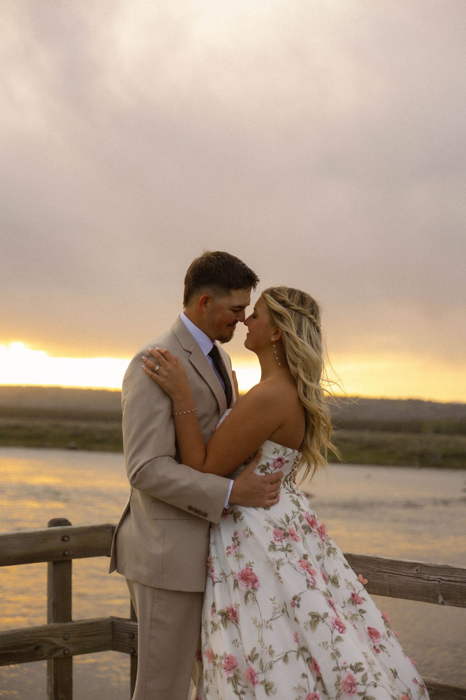 Couple sharing a romantic moment at sunset on a wooden bridge, man in tan suit, woman in floral dress.