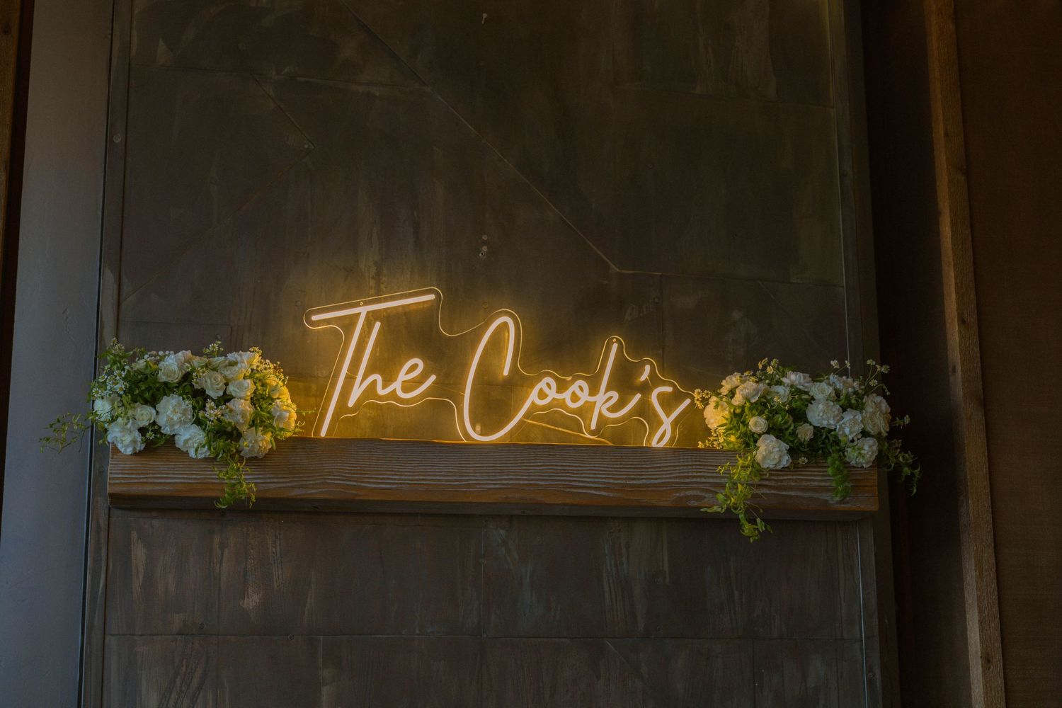 Golden neon sign reading The Cook's displayed on a wooden shelf with white and yellow floral arrangements.