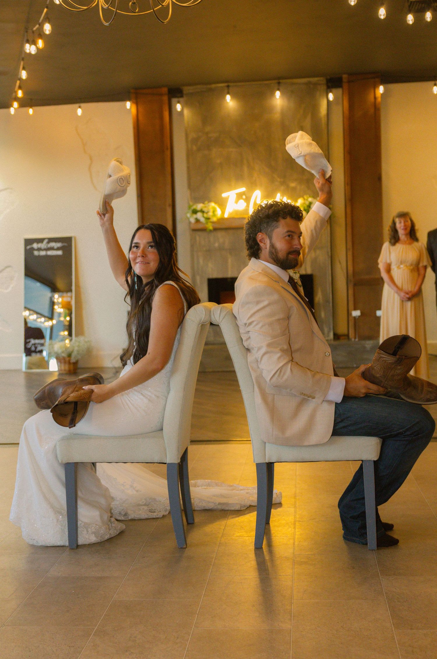 Bride and groom sit back-to-back on elegant chairs, playfully holding shoes aloft during a wedding reception game.