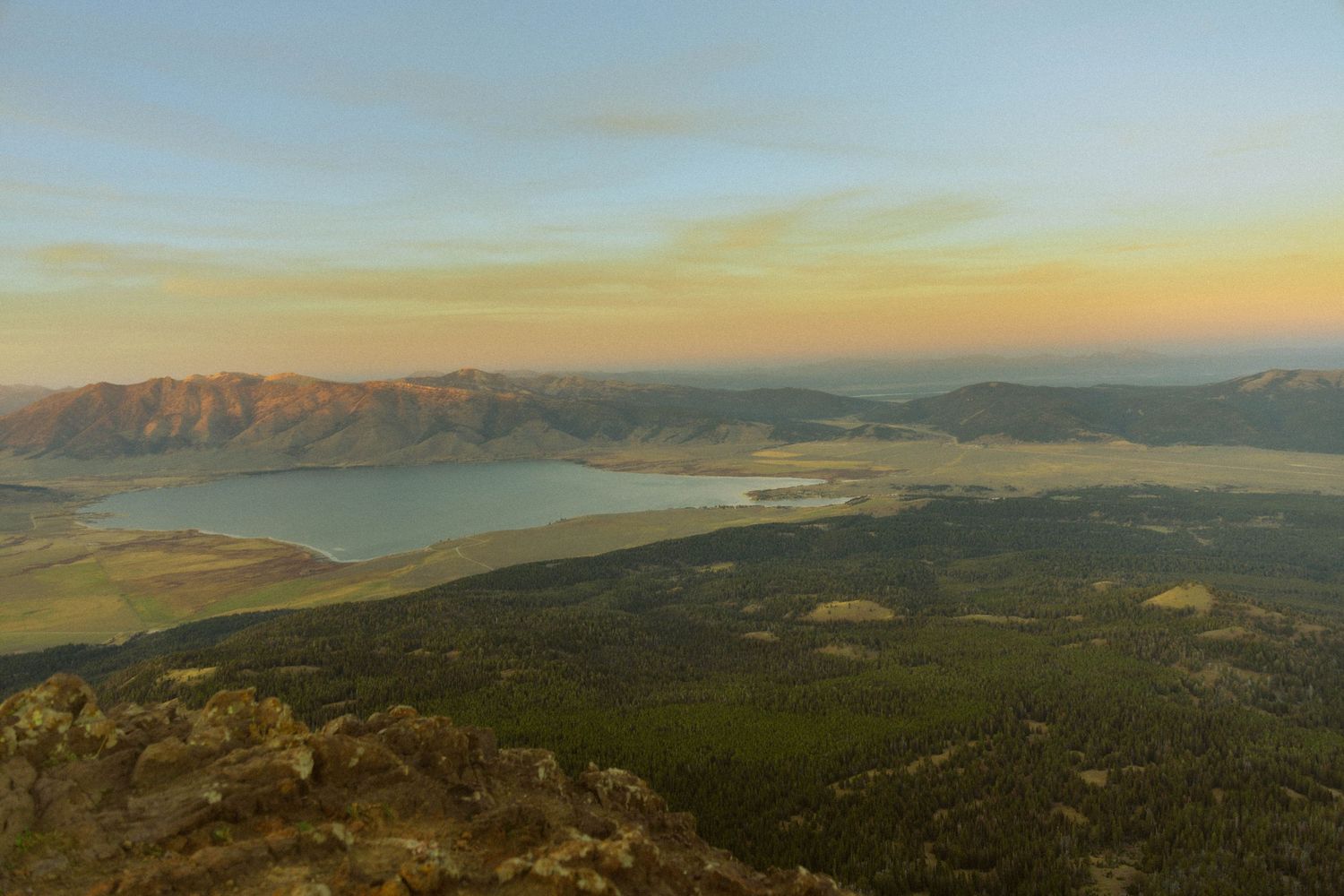 Aerial sunset view of a vast mountain lake valley with golden skies, rolling hills, and lush green forests in the foreground.