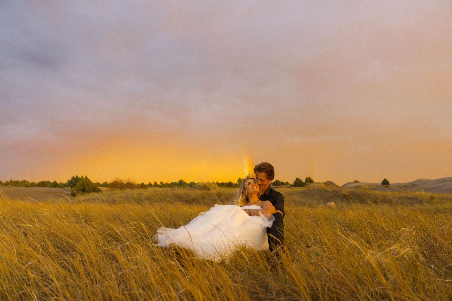 Groom dips bride in golden wheat field at sunset with rainbow sky backdrop during outdoor wedding photo session.
