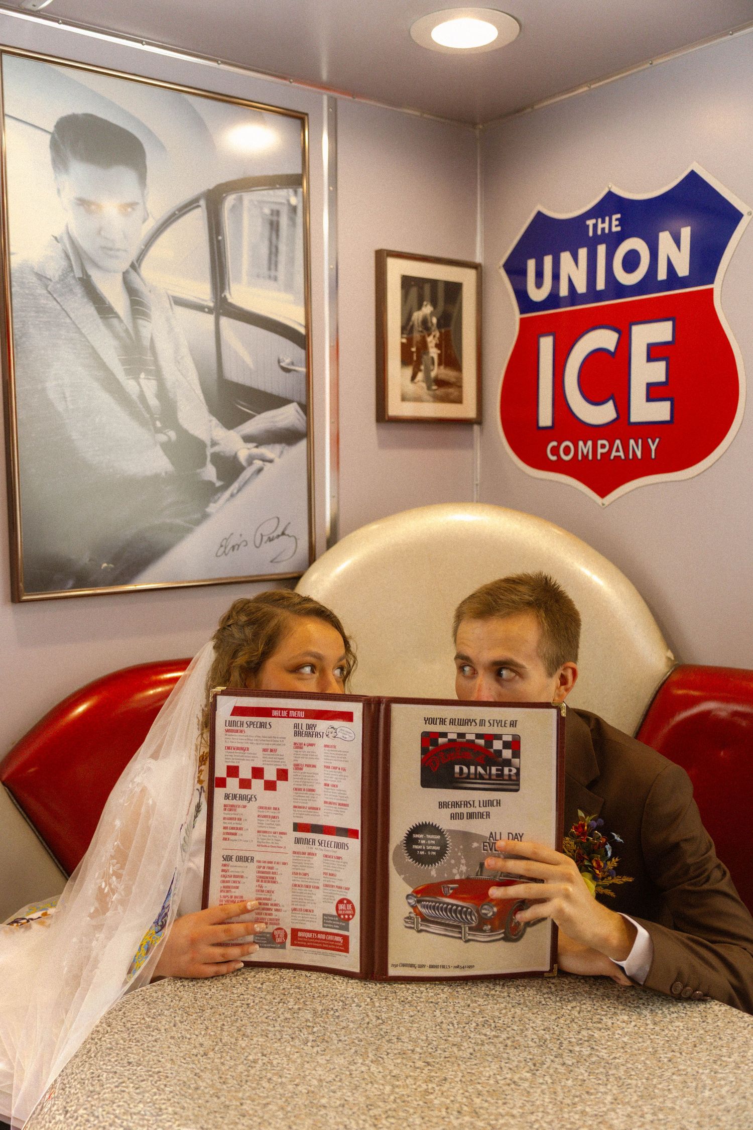 Newlywed couple hiding behind a diner menu at a retro booth with Elvis poster and Union Ice Company sign.