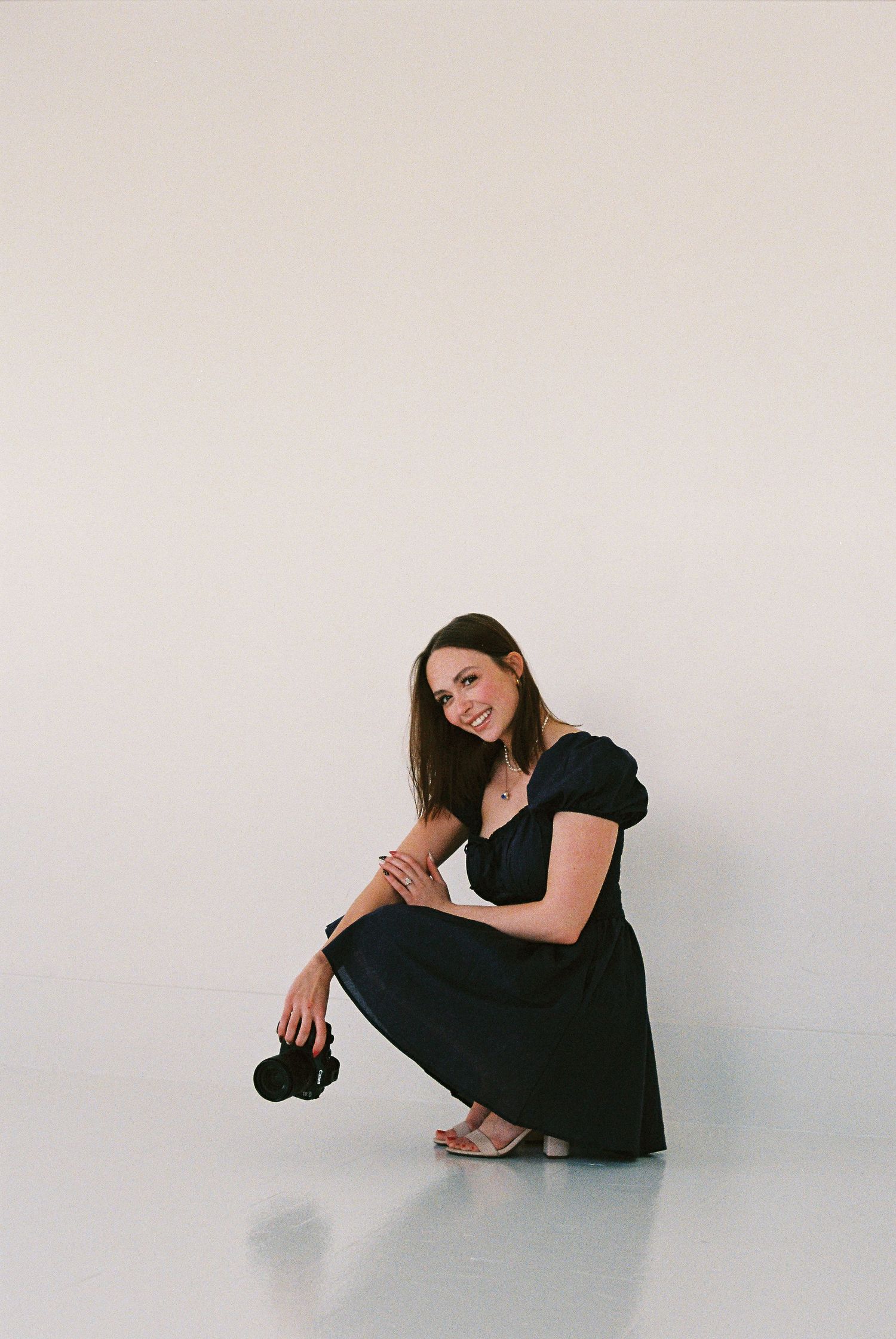 Woman in black dress crouching while holding a camera, smiling in a minimalist white studio setting.