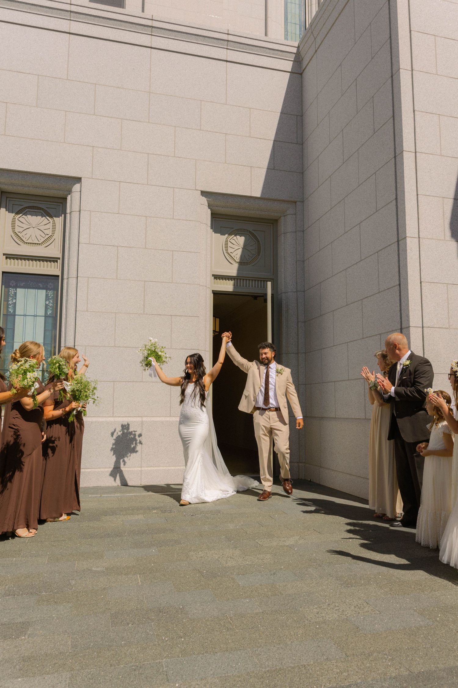 Newlywed couple exits wedding venue raising hands, surrounded by bridesmaids in brown dresses and guests applauding.