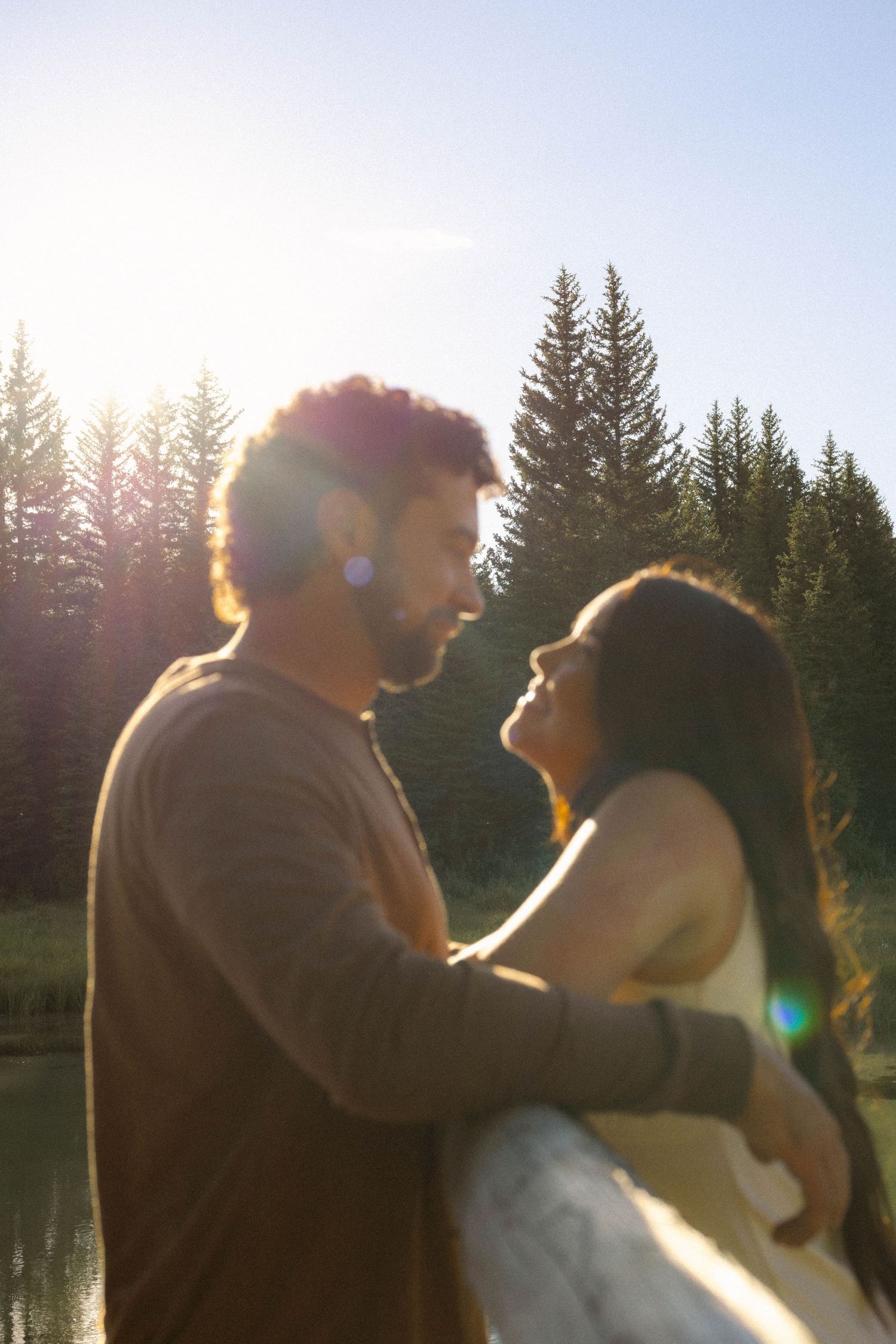 Couple embracing near a sunlit forest lake, sharing a romantic moment with pine trees and lens flare in background.