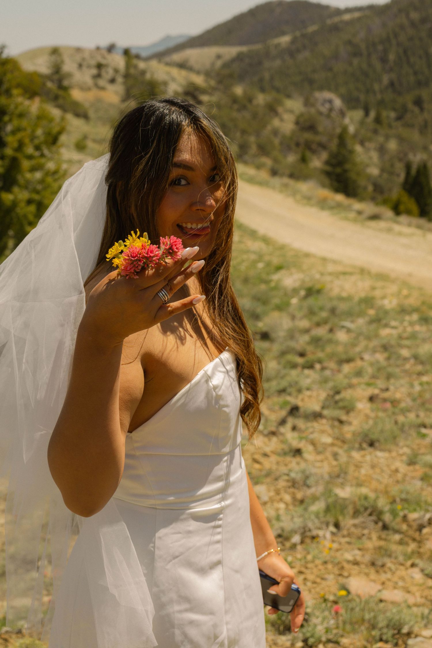 Smiling bride in white strapless gown and veil holds wildflowers outdoors with scenic mountain landscape behind her.