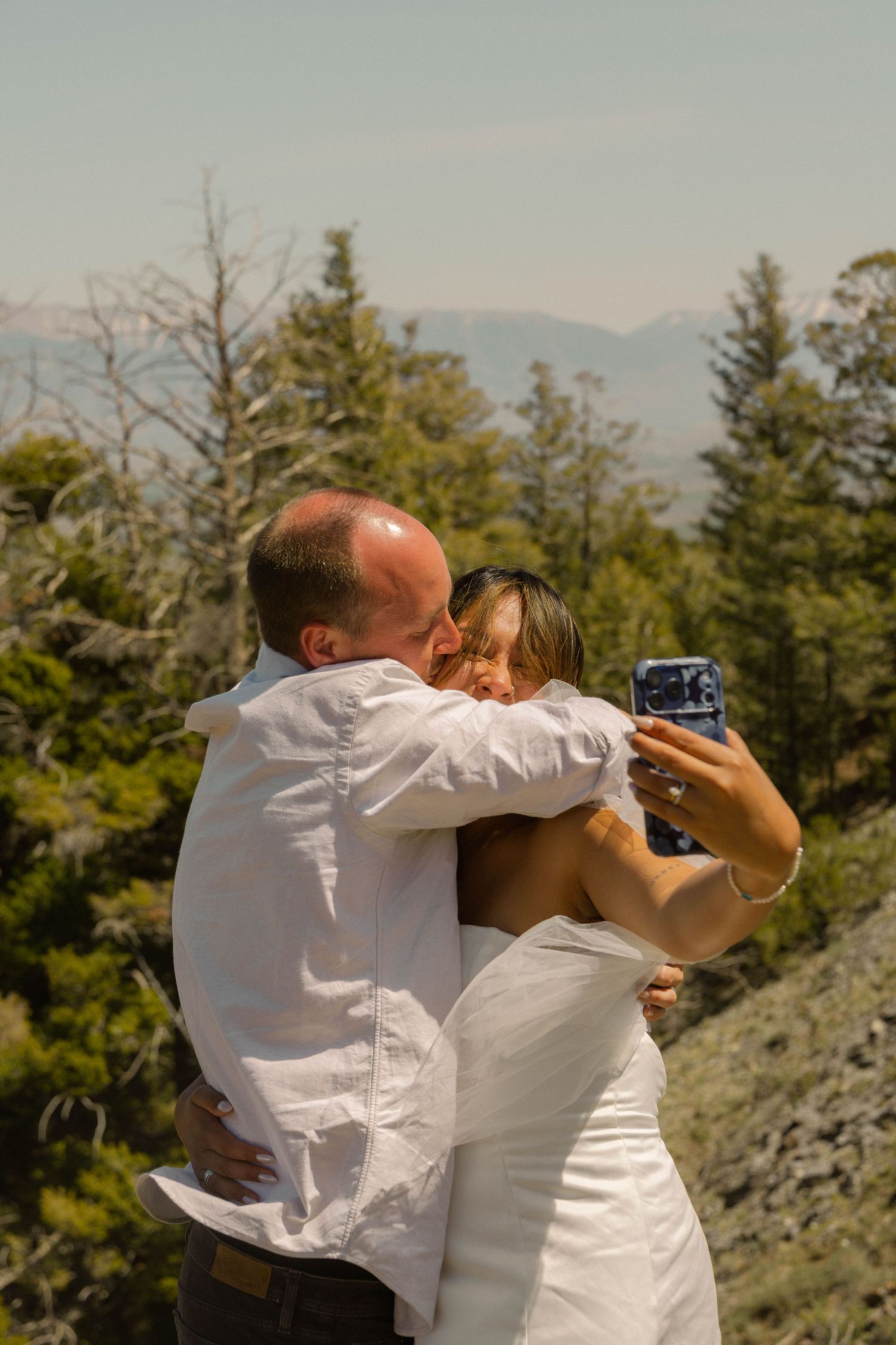 Couple in white attire embracing and taking a selfie outdoors with mountain and pine tree backdrop.