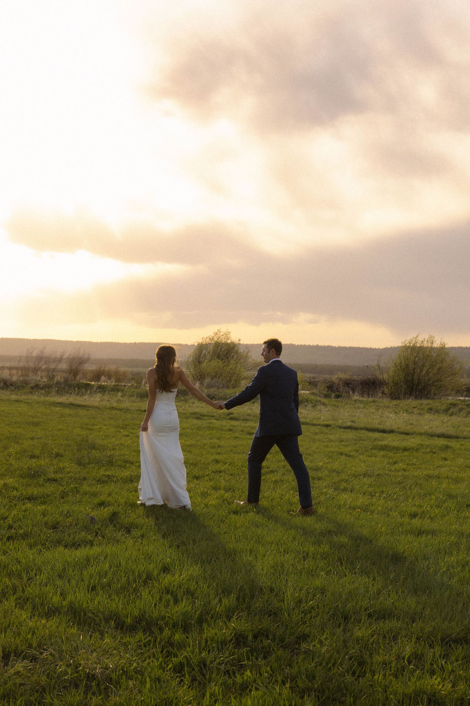 Bride and groom holding hands in a green field at golden sunset, walking together in wedding attire.