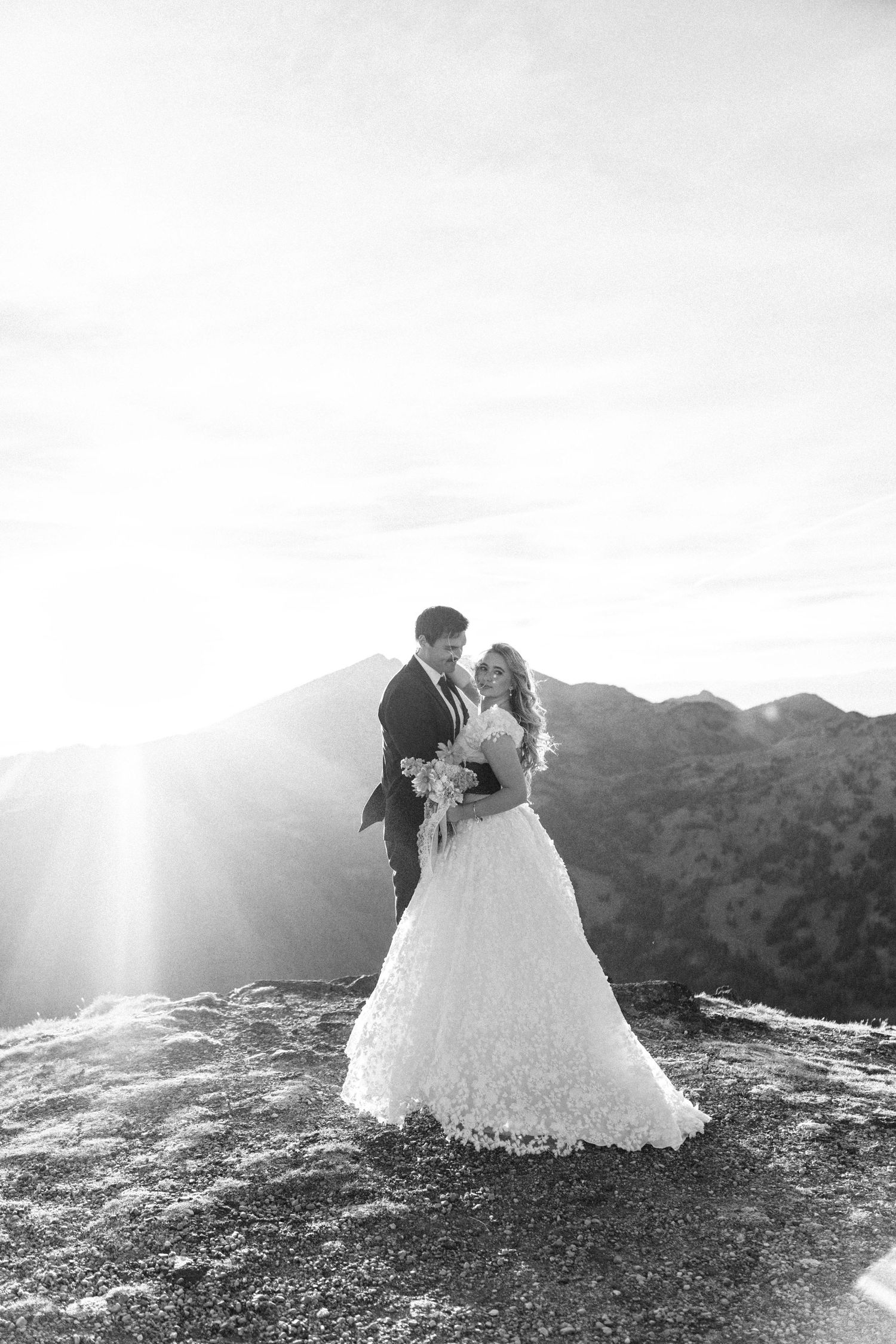 Black and white photo of bride and groom embracing on mountain summit with dramatic sunburst and scenic peaks behind them.