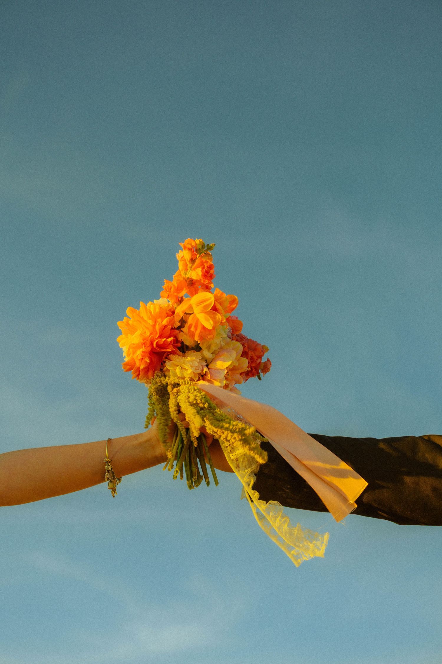Two hands holding a vibrant orange and yellow flower bouquet with ribbon against a clear blue sky.