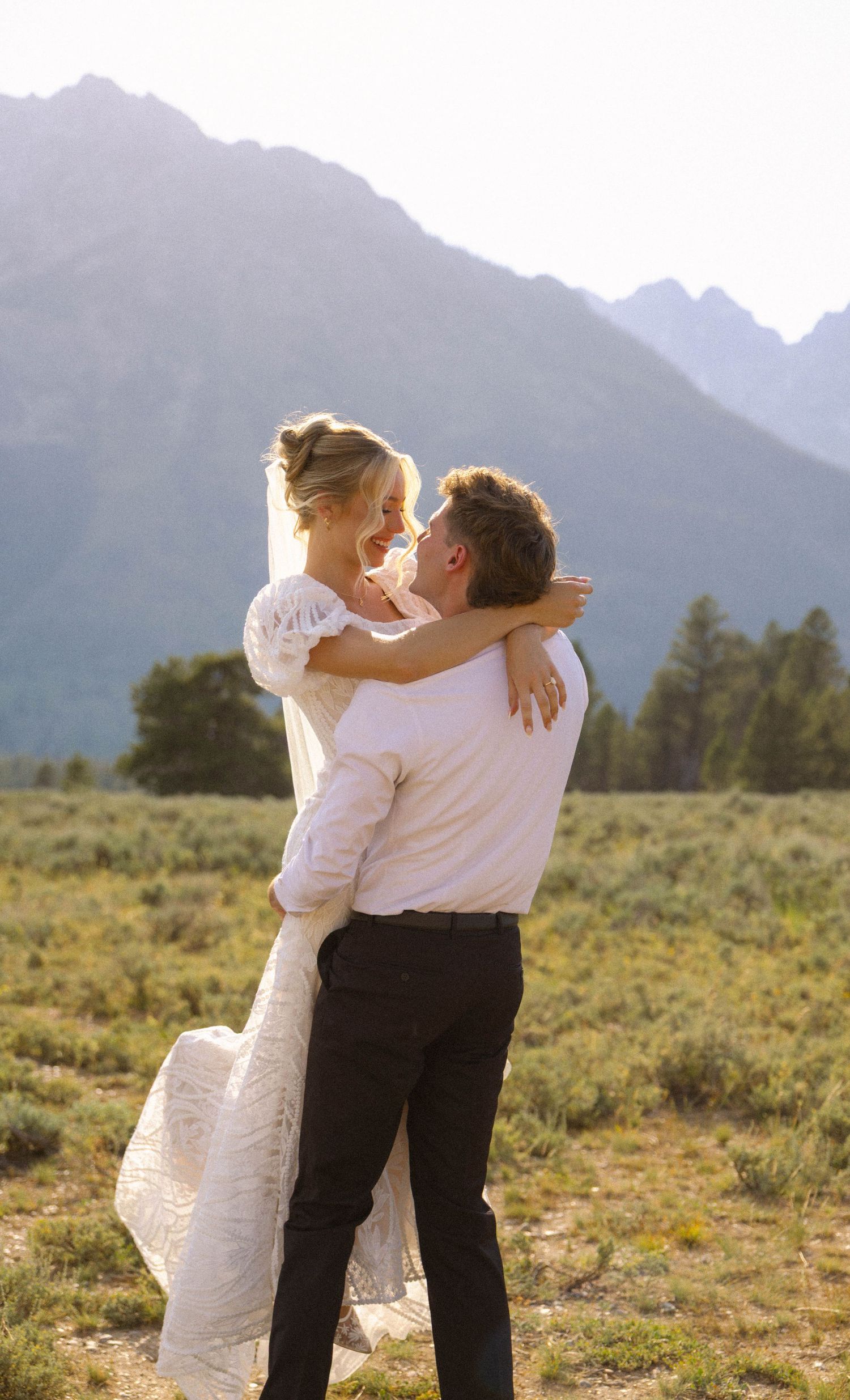 A groom lifts his bride in a white wedding dress, sharing a joyful embrace with mountain scenery behind them.