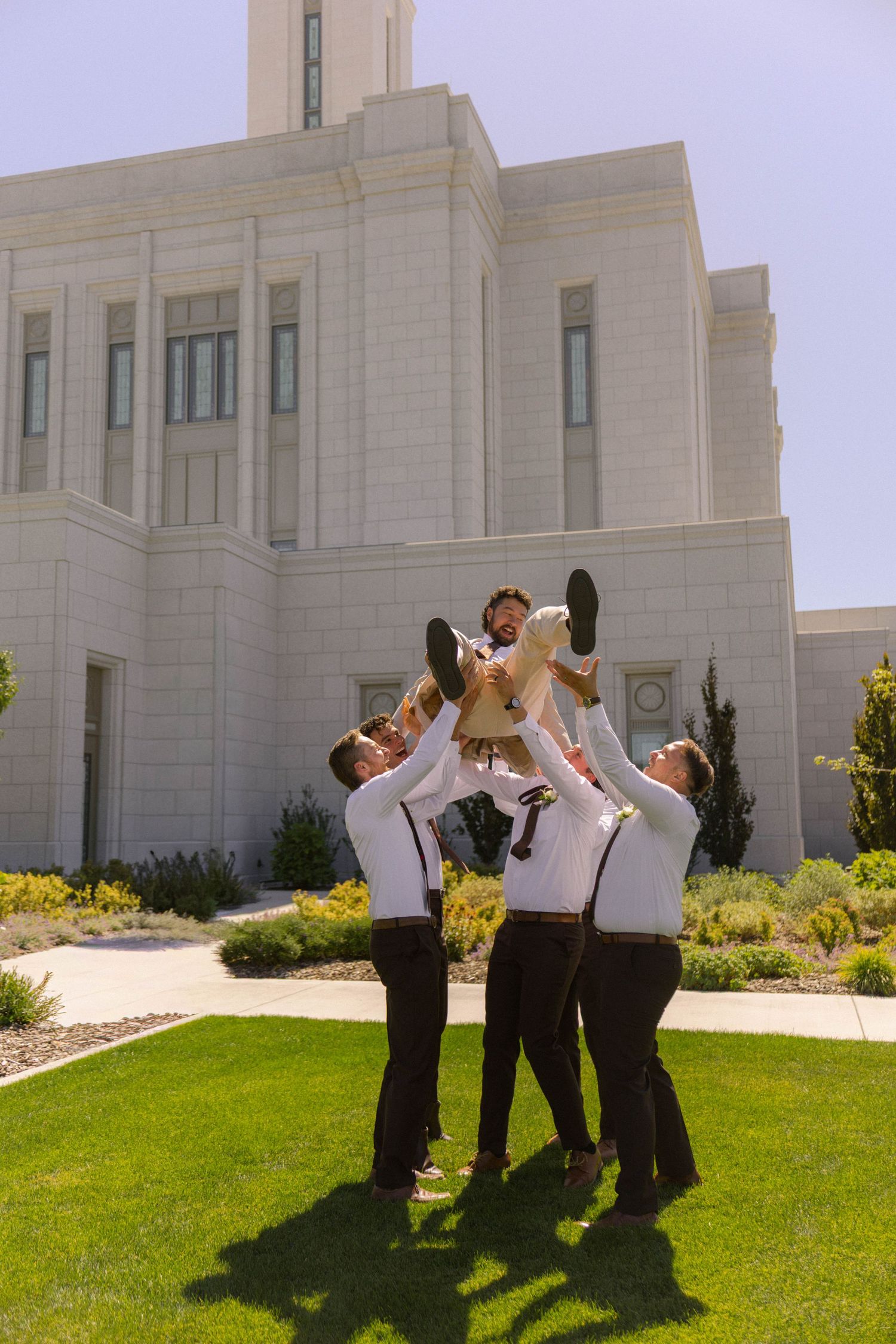 Groomsmen lifting groom in front of a white stone temple on a sunny wedding day.