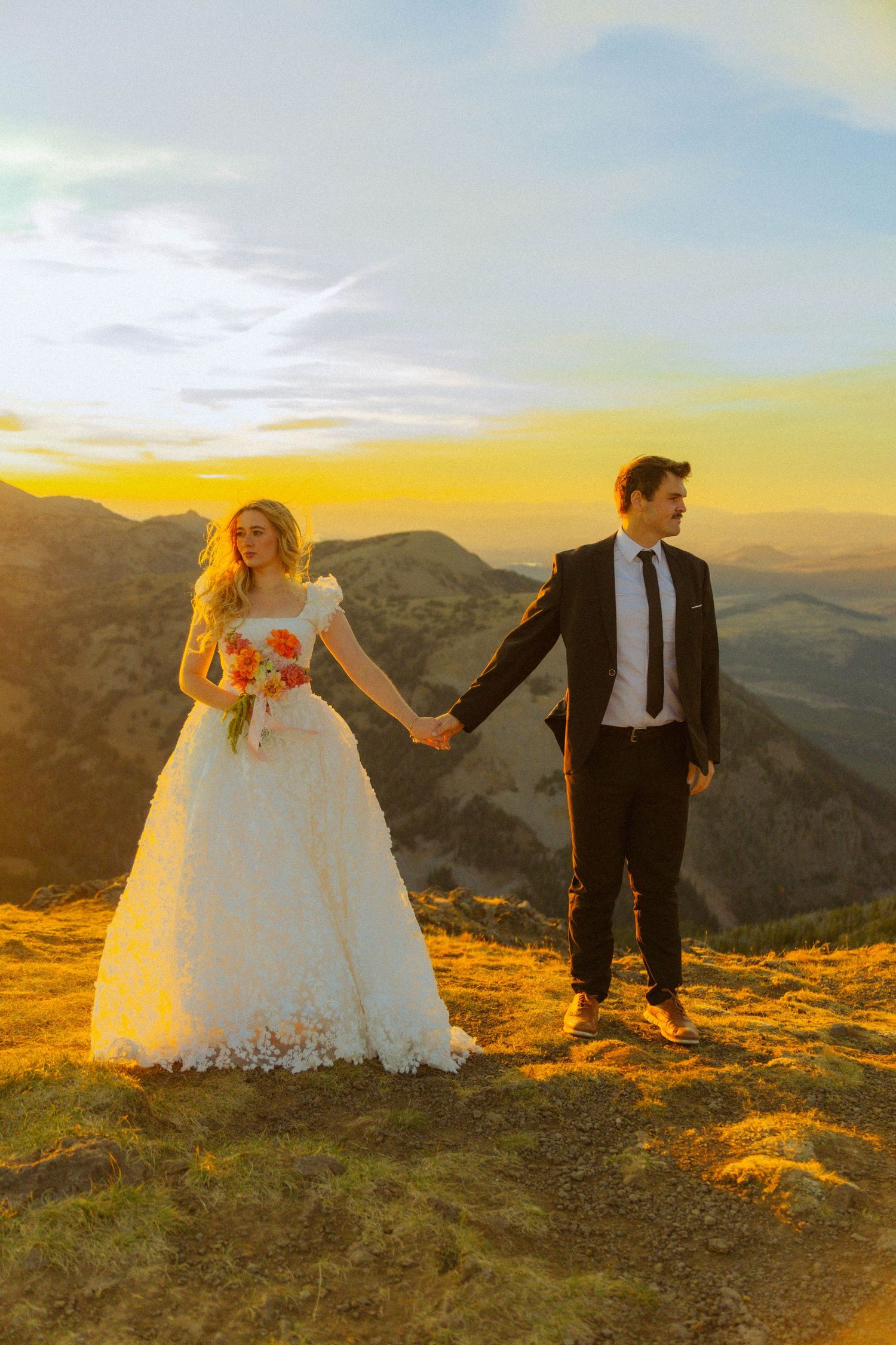 Bride and groom hold hands at golden sunset on mountain peak, bride in floral white gown holding bouquet.
