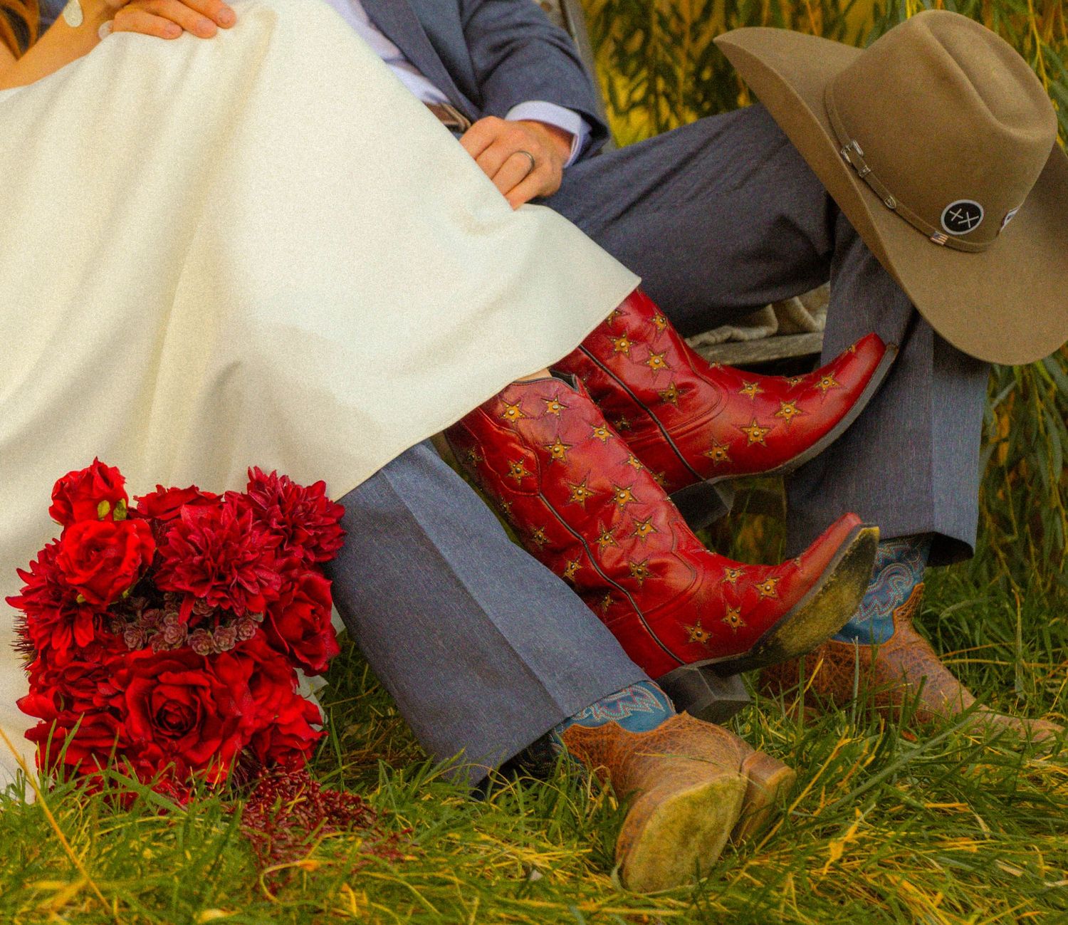 Bride in white dress with red cowboy boots and bouquet beside groom in gray suit and cowboy hat outdoors.