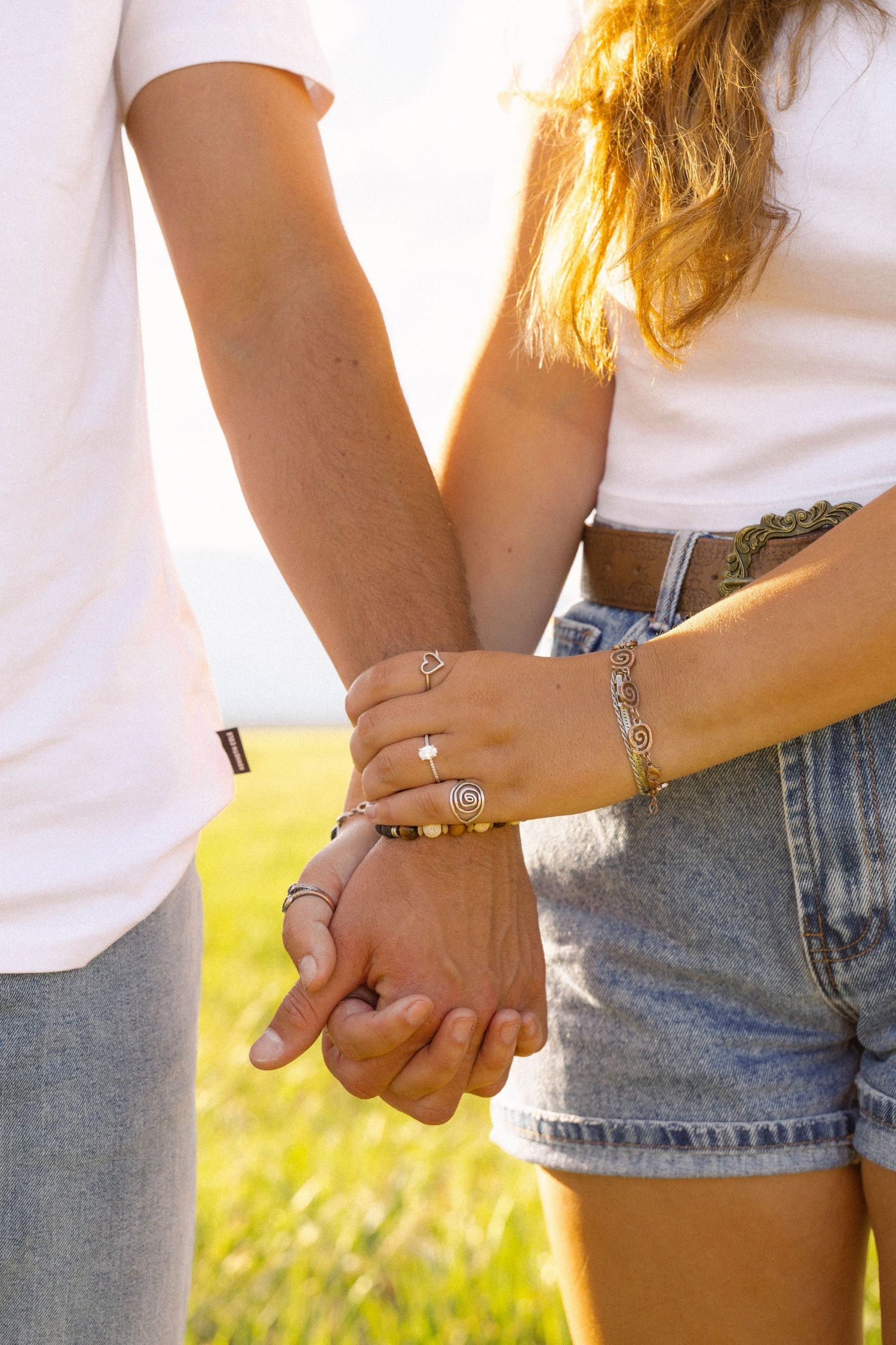 Couple holding hands outdoors at golden hour, woman wearing rings and bracelets, both in white tees and denim.