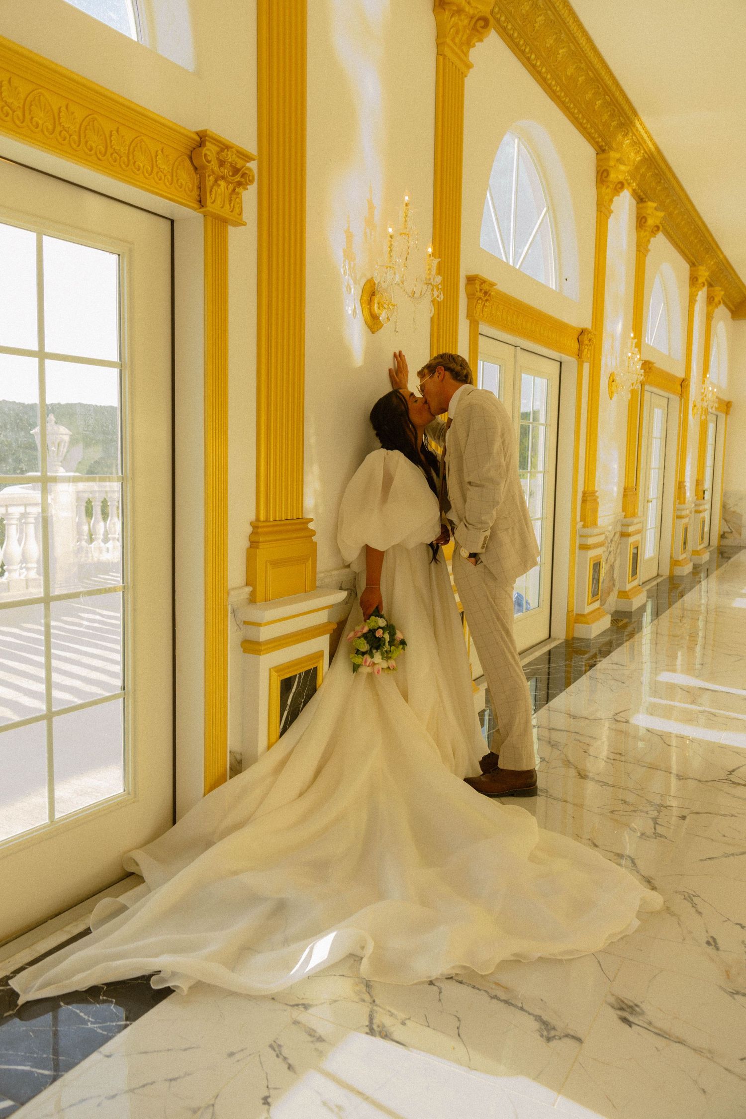 Bride and groom kissing in elegant gold and marble hallway, bride wearing flowing white gown with long train.