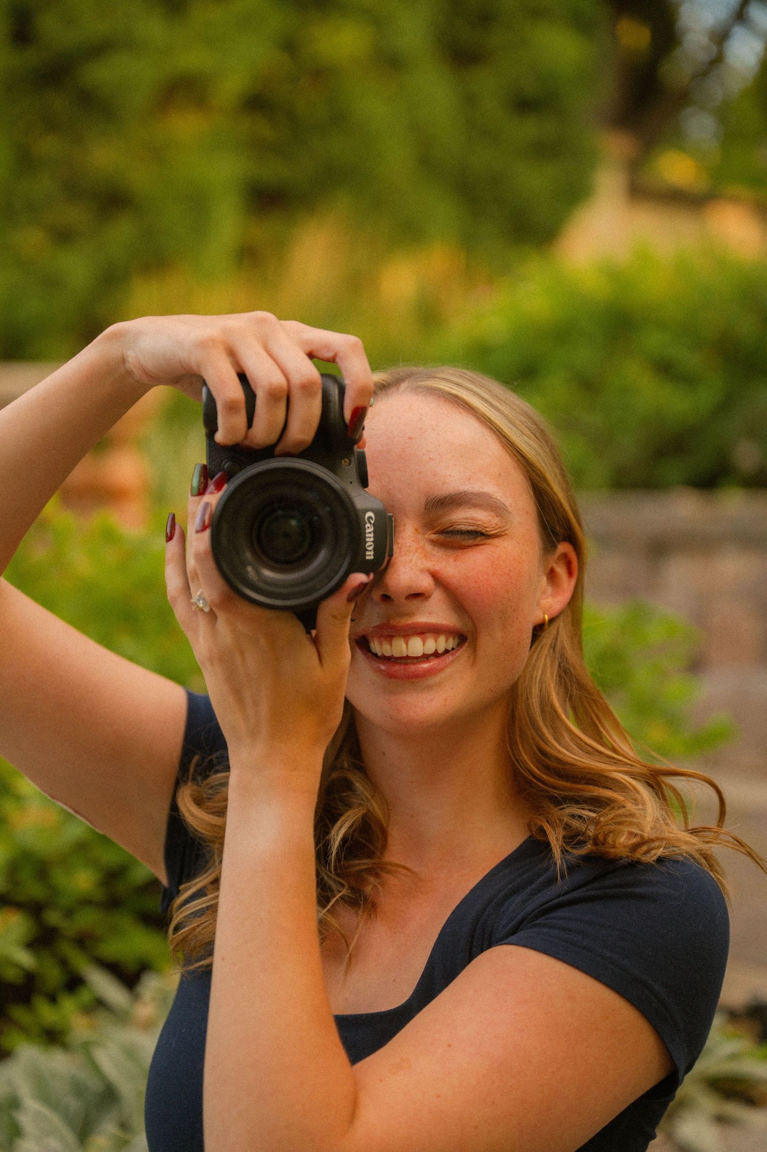 Smiling blonde woman holding a Canon DSLR camera up to her eye outdoors in a lush green garden setting.