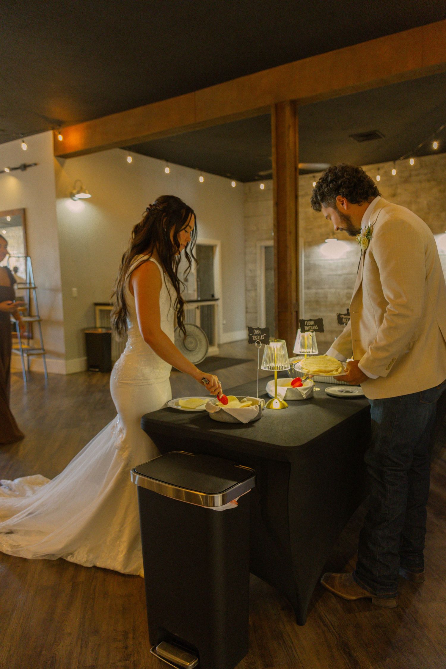 Bride and groom cutting wedding cake at a rustic indoor venue with string lights and wooden beams.