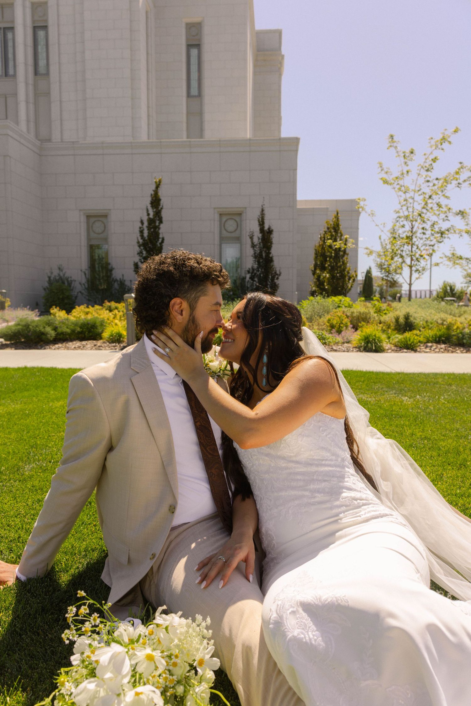 A bride and groom share a romantic moment sitting on green grass outside a white temple on their wedding day.