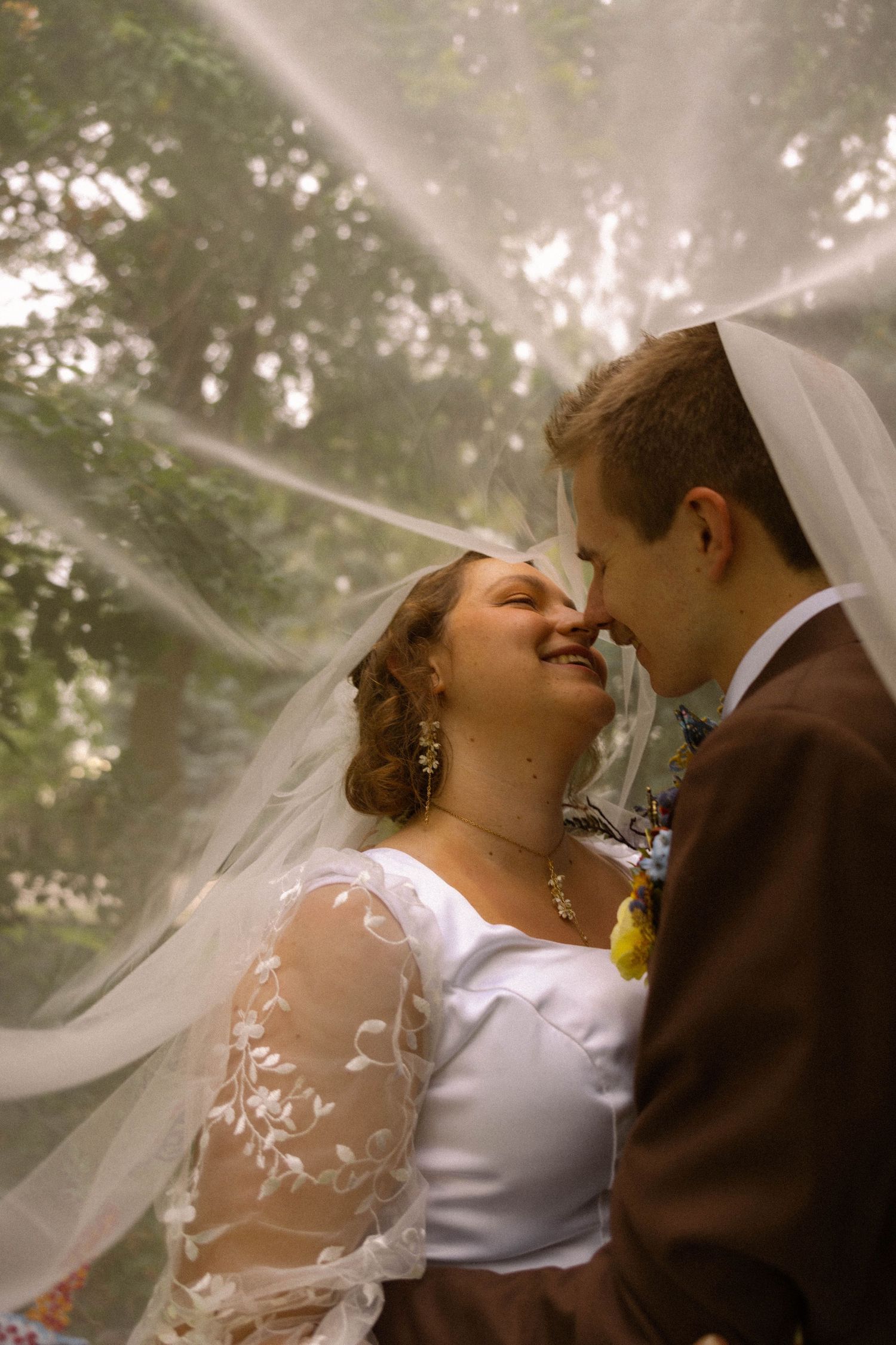 Bride and groom share a romantic moment under a flowing veil outdoors, smiling and touching noses on their wedding day.