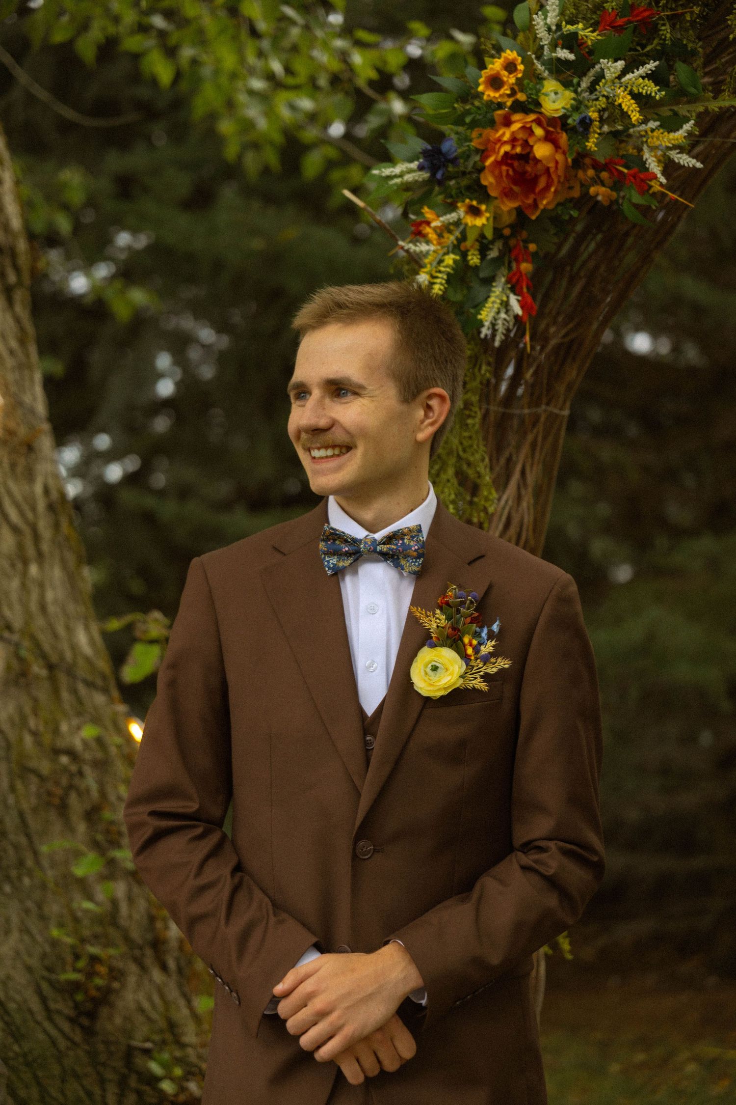 Smiling groom in brown suit with floral boutonniere and bow tie stands outdoors near colorful wedding arch.